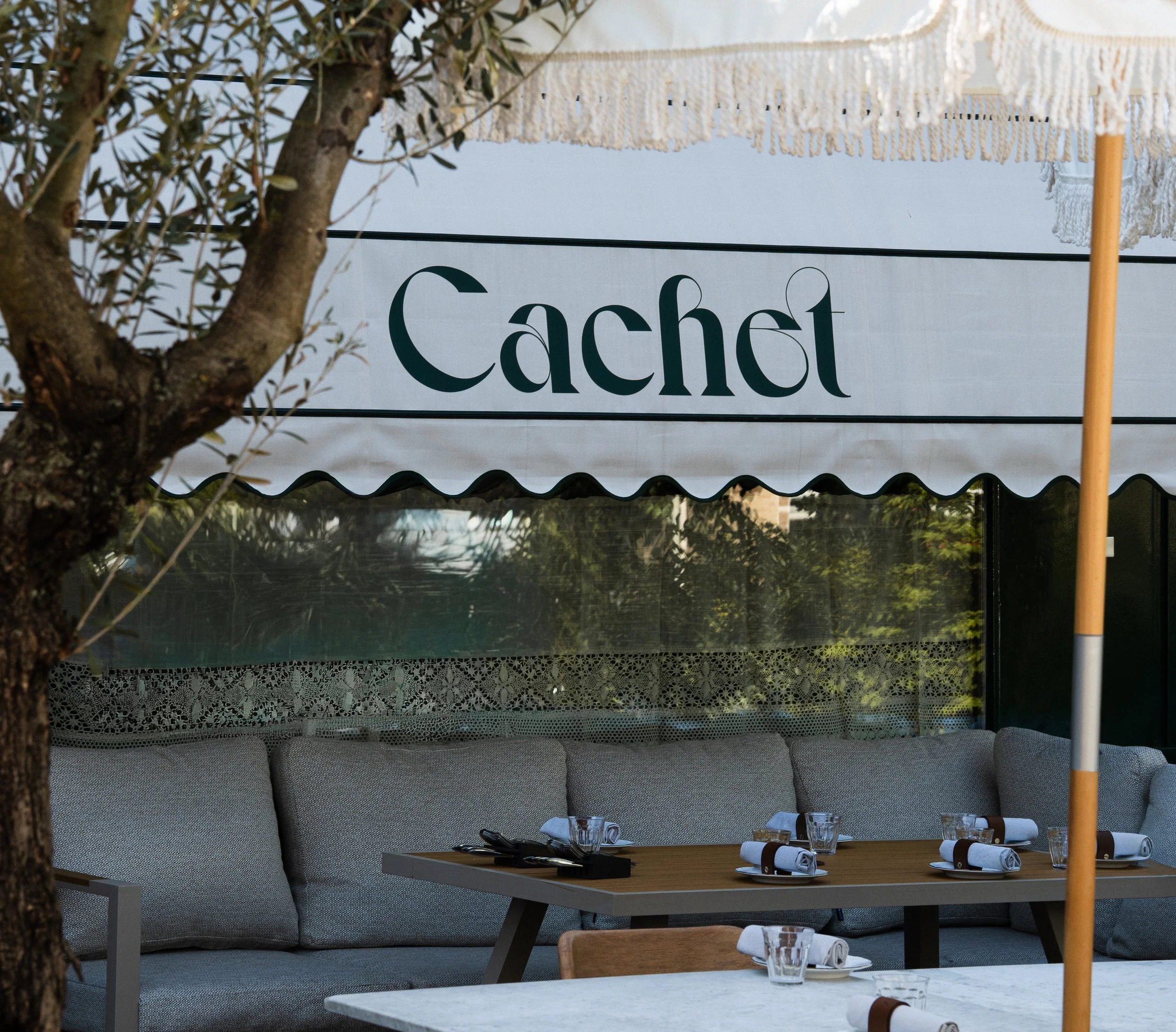 Een restaurant of café binnen- of buitenruimte met een tafel die is gedekt met glaswerk, servetten en bestek. Op de achtergrond hangt een doek met de tekst 'Caché' en zitgelegenheid met kussens.