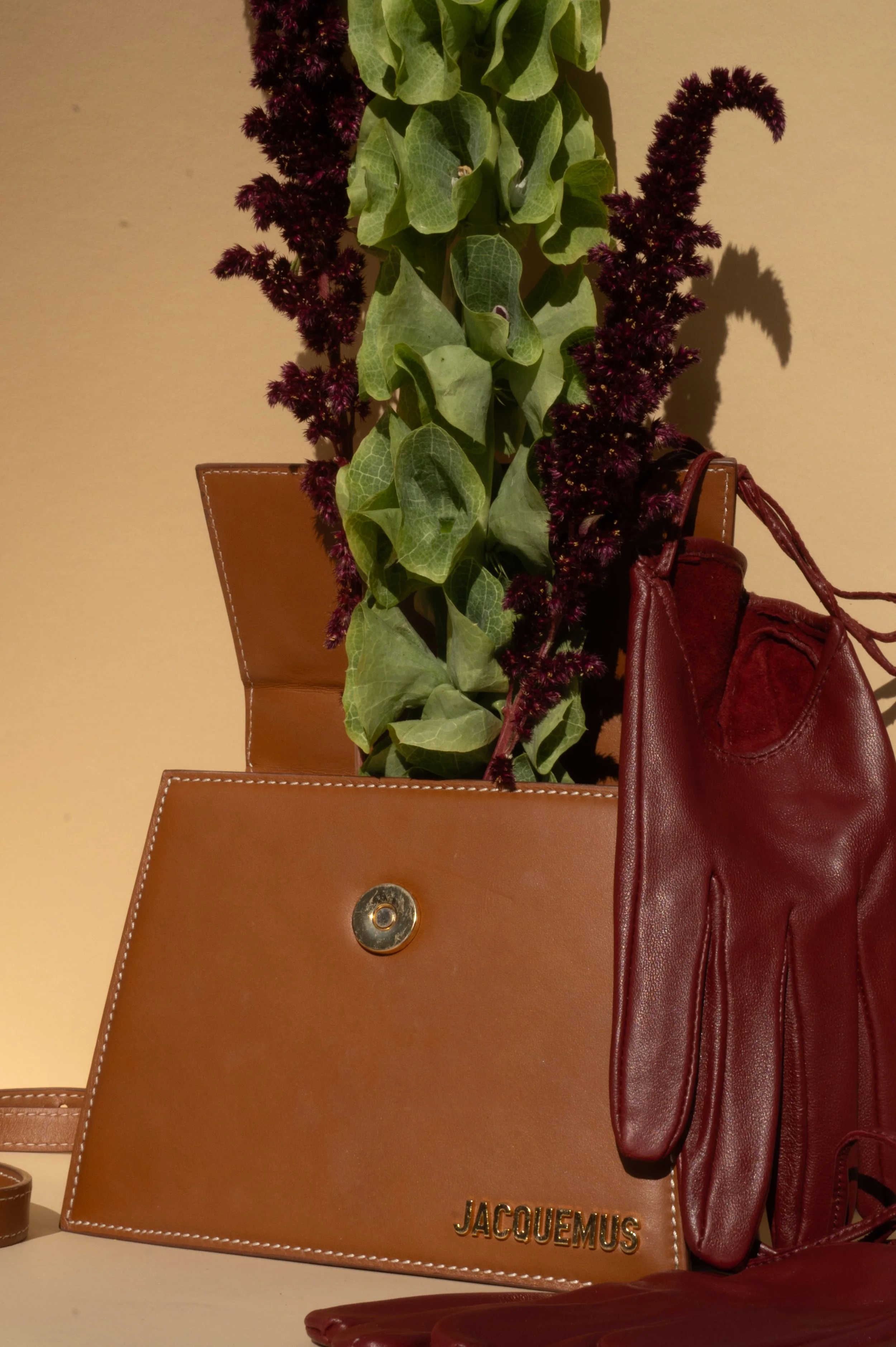 Een bloemstuk met groene bladeren en paarse bloemen, geplaatst in een set van drie tassen, waaronder een lichtbruine Jacquemus clutch en een rode leren tas.
