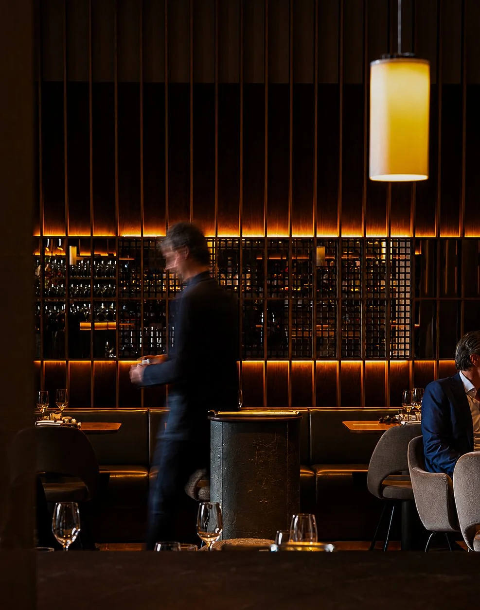 Binnen een restaurant met een houten wand en warme verlichting, een ober die loopt, en gasten die aan tafels zitten.