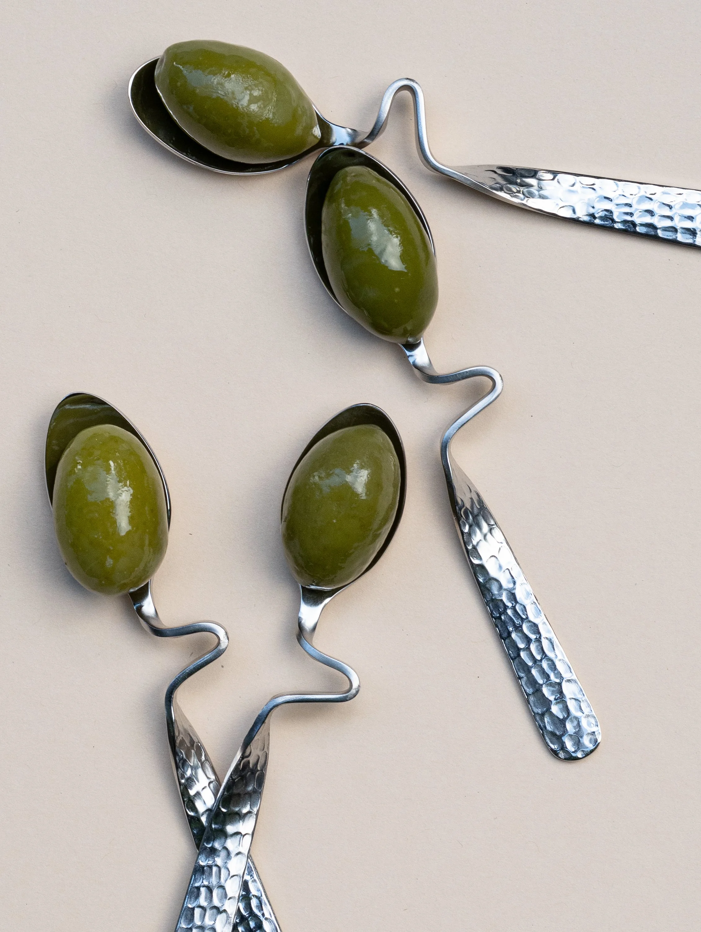Drie eetlepels met groene olijven met metalen handvatten, uitgestald op een lichtgekleurde achtergrond.