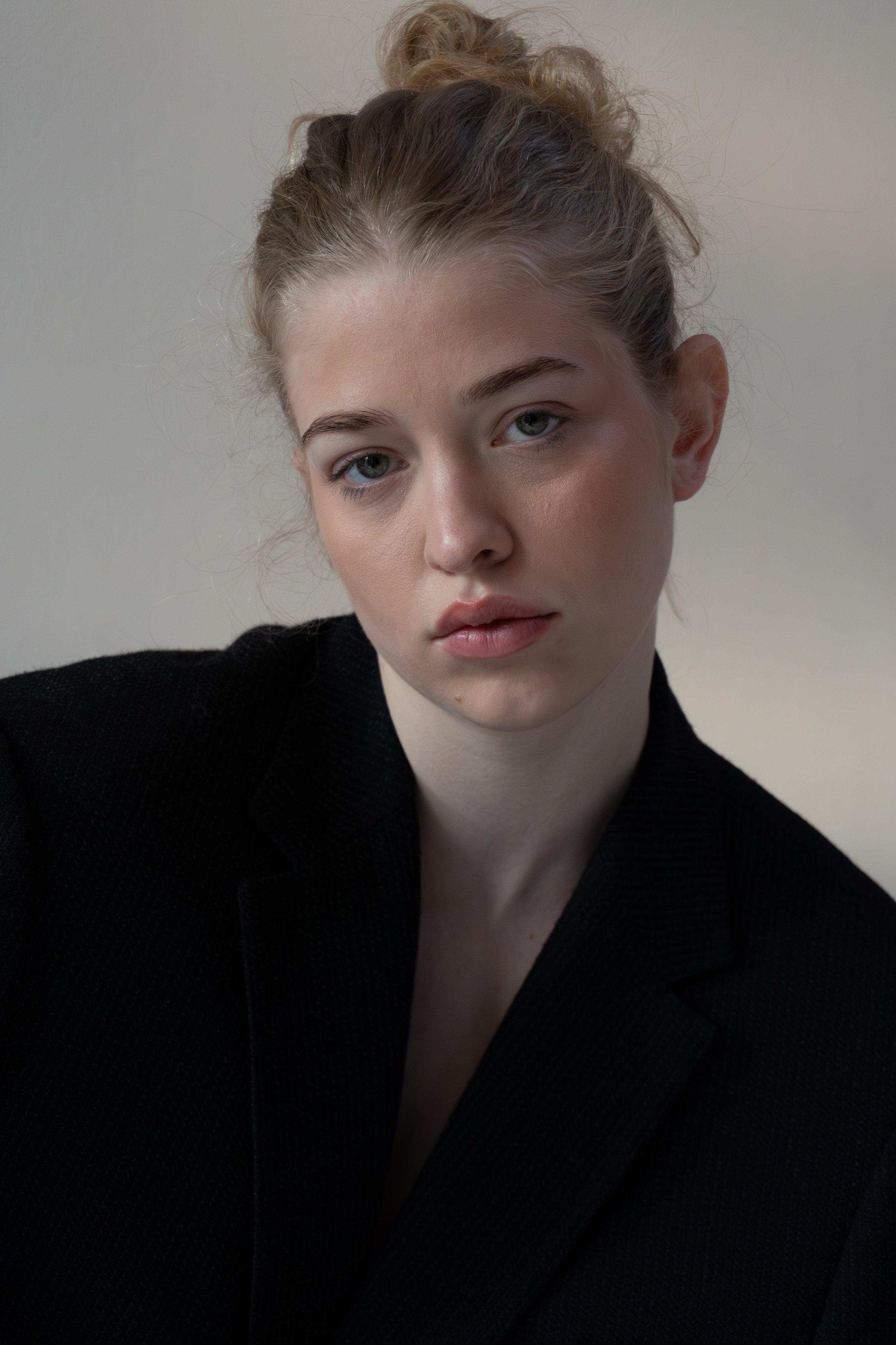 Close-up van een jonge vrouw met lichtbruine haar in een rommelige knot, frisse make-up, blauwe ogen, in een zwarte blouse, in een neutrale achtergrond.