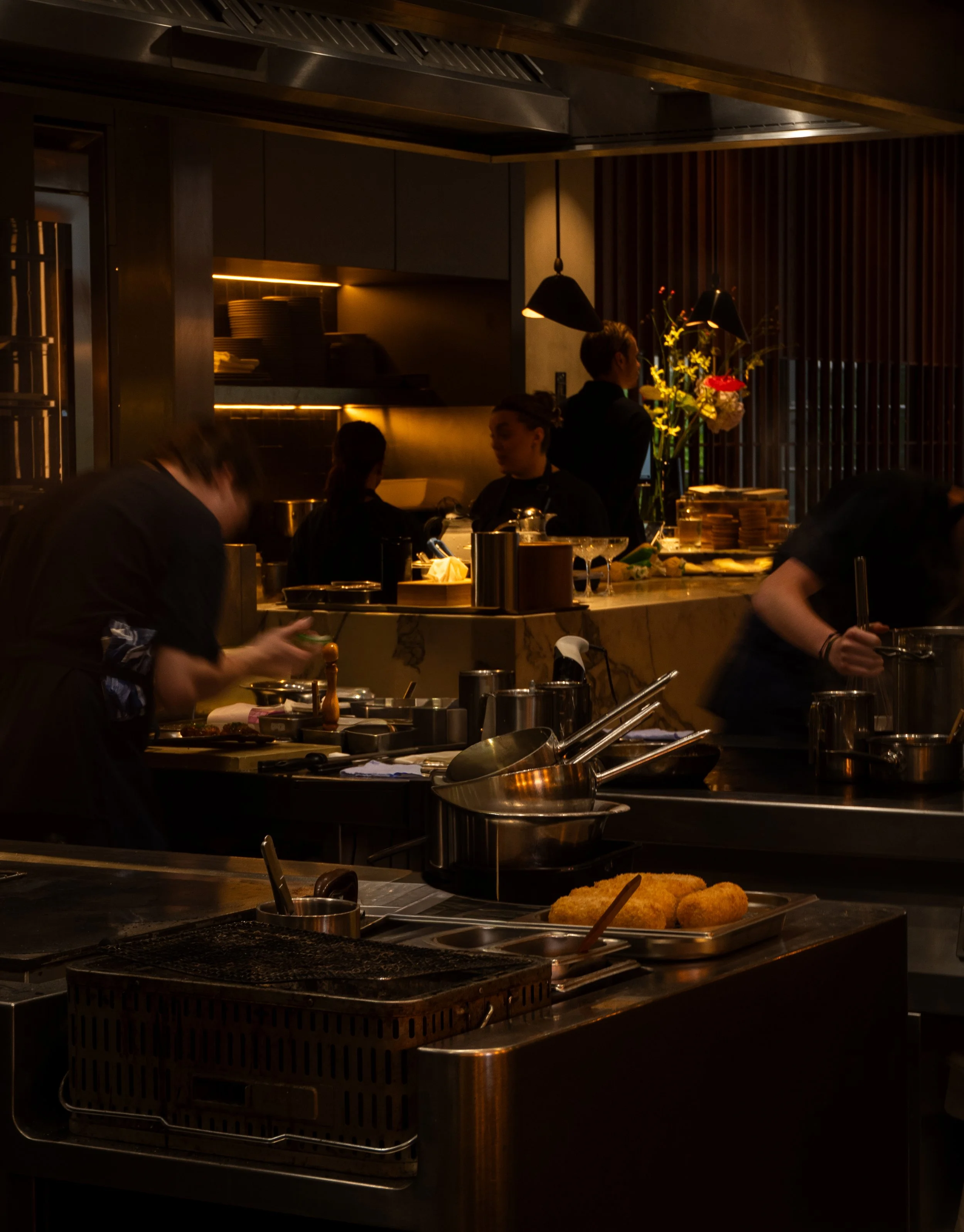 Een restaurantkeuken waar koks gerechten bereiden. In de voorgrond liggen gefrituurde kroketten op een bord, op de achtergrond zijn mensen bezig met koken en serveren.