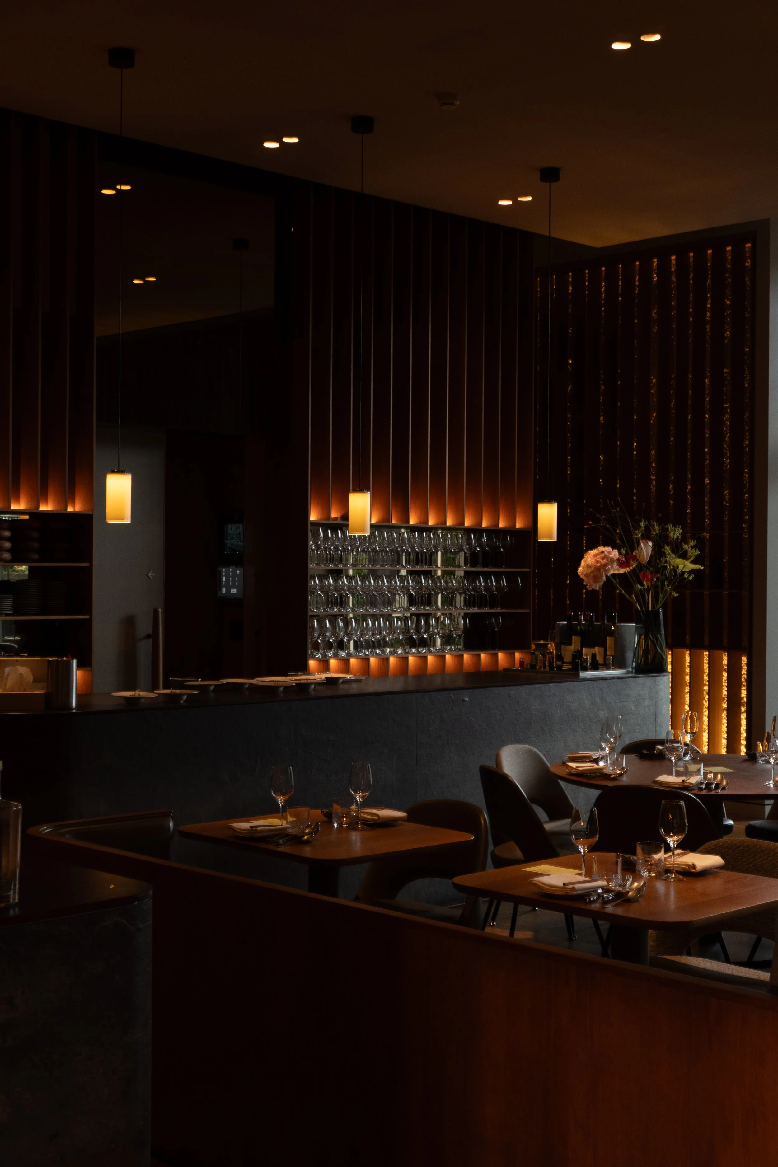 Elegant restaurant met gedimde verlichting, bloemen op de tafel, glazen en borden klaar voor gasten, en een decoratieve houten scheidingswand.