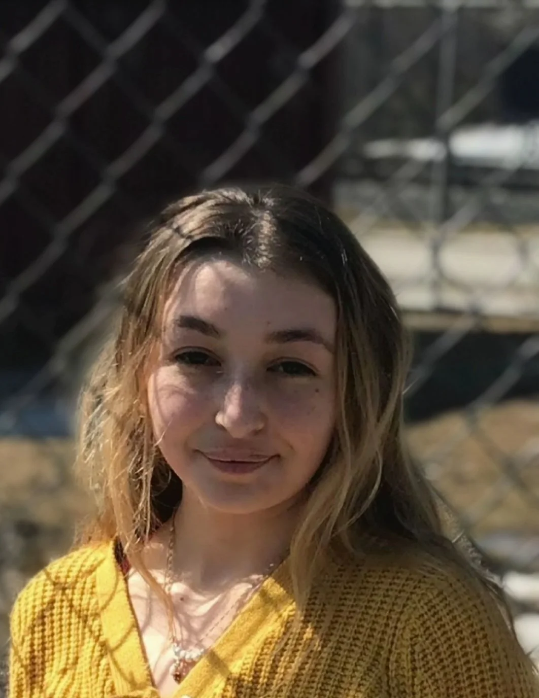 A young woman with wavy, shoulder-length hair wearing a yellow sweater, standing outdoors near a chain-link fence.