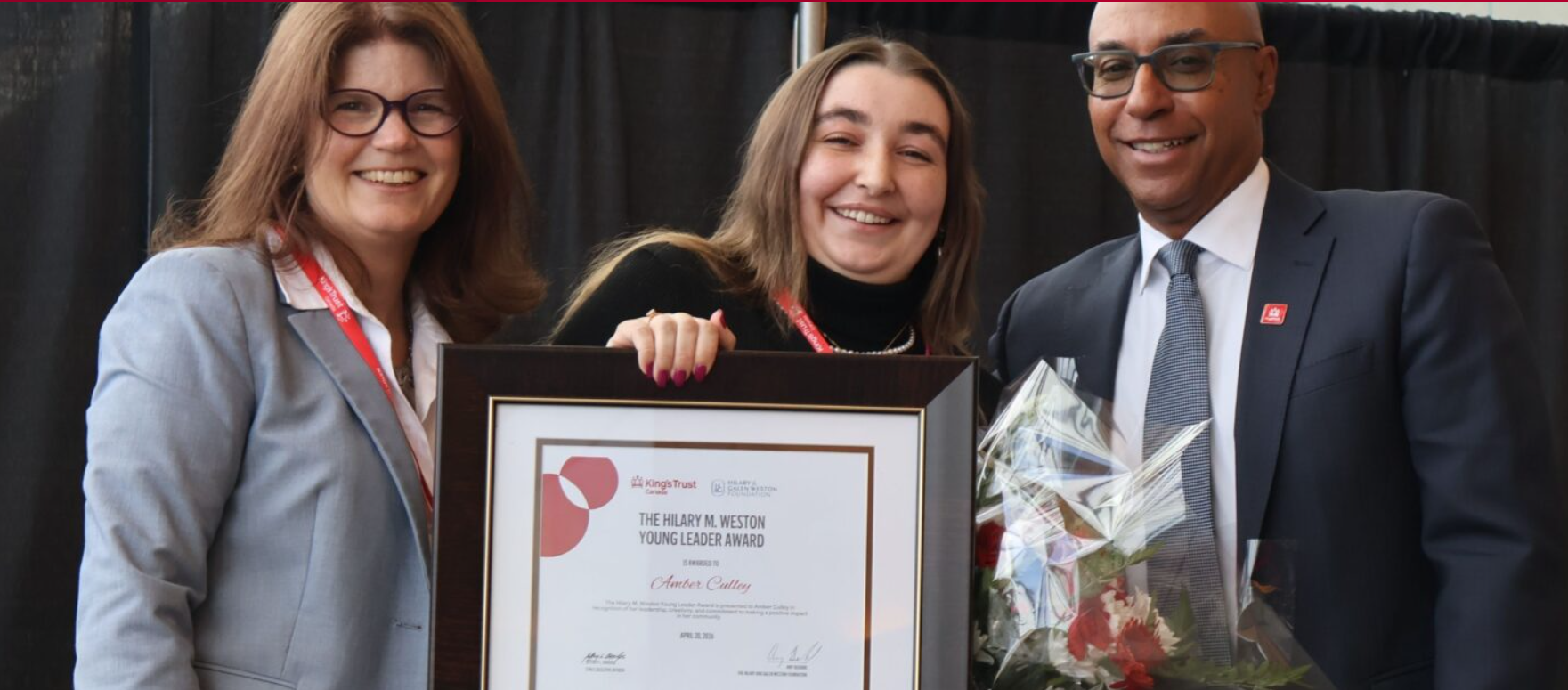 Three people smiling at an award ceremony, with one holding a framed award certificate and flowers. The person in the middle is a young woman, and the two others are adults dressed in business attire.