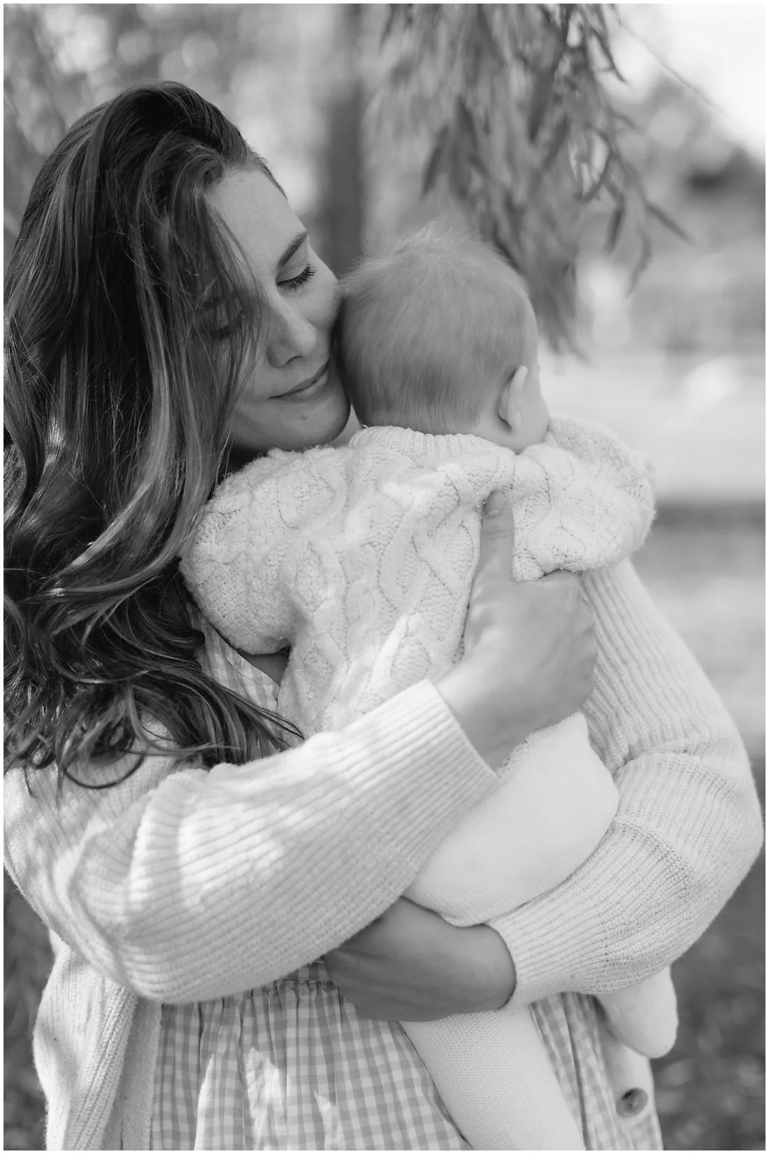 A woman with long wavy hair holding a young child close to her chest. The woman has her eyes closed and is gently smiling, while the child is facing away from the camera and holding onto her. They are outdoors with blurred trees in the background.