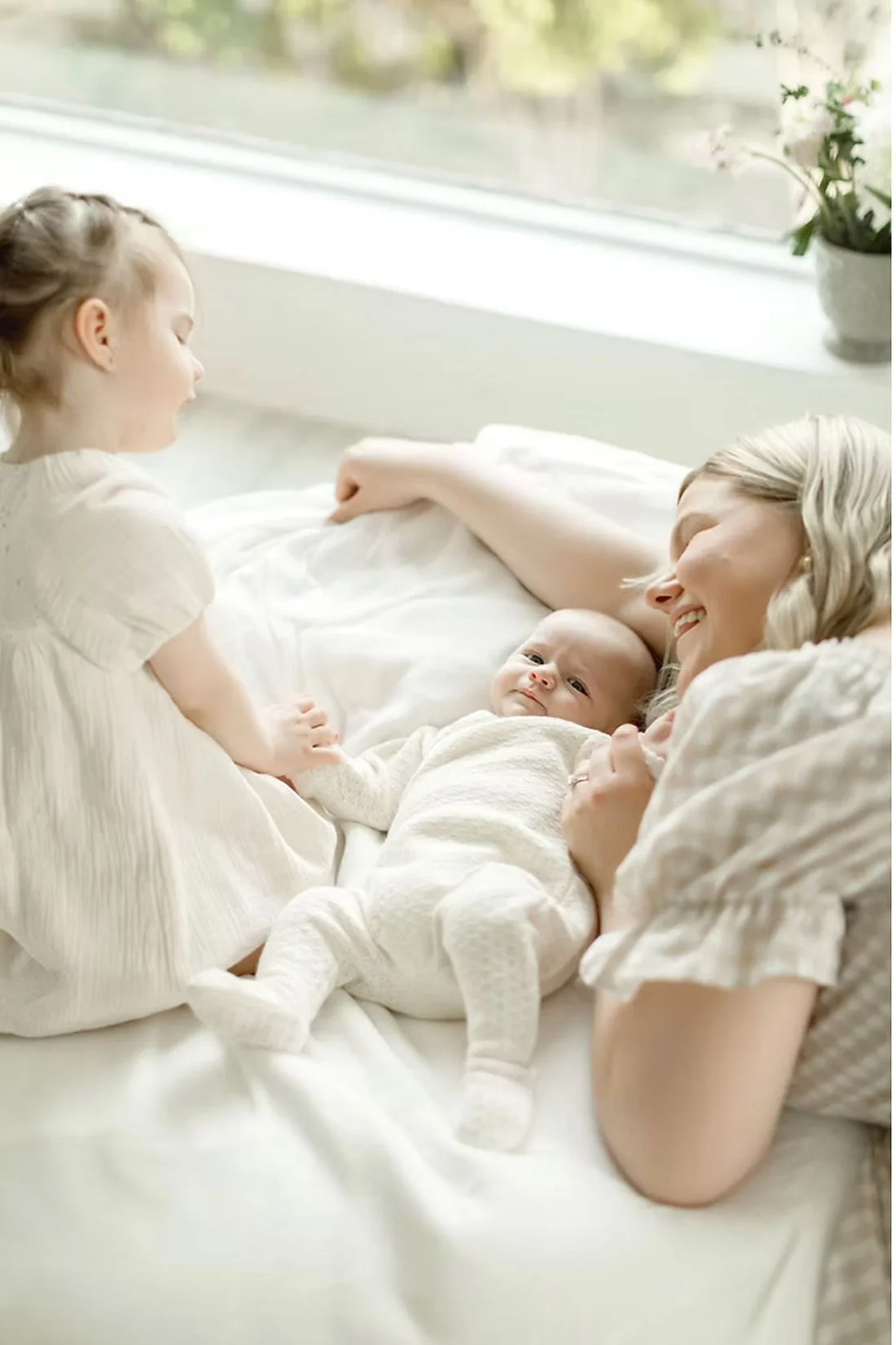 A woman with gray hair lying on a bed with a baby in her arms, and a young girl sitting beside them, all smiling and interacting indoors near a window with a potted plant.
