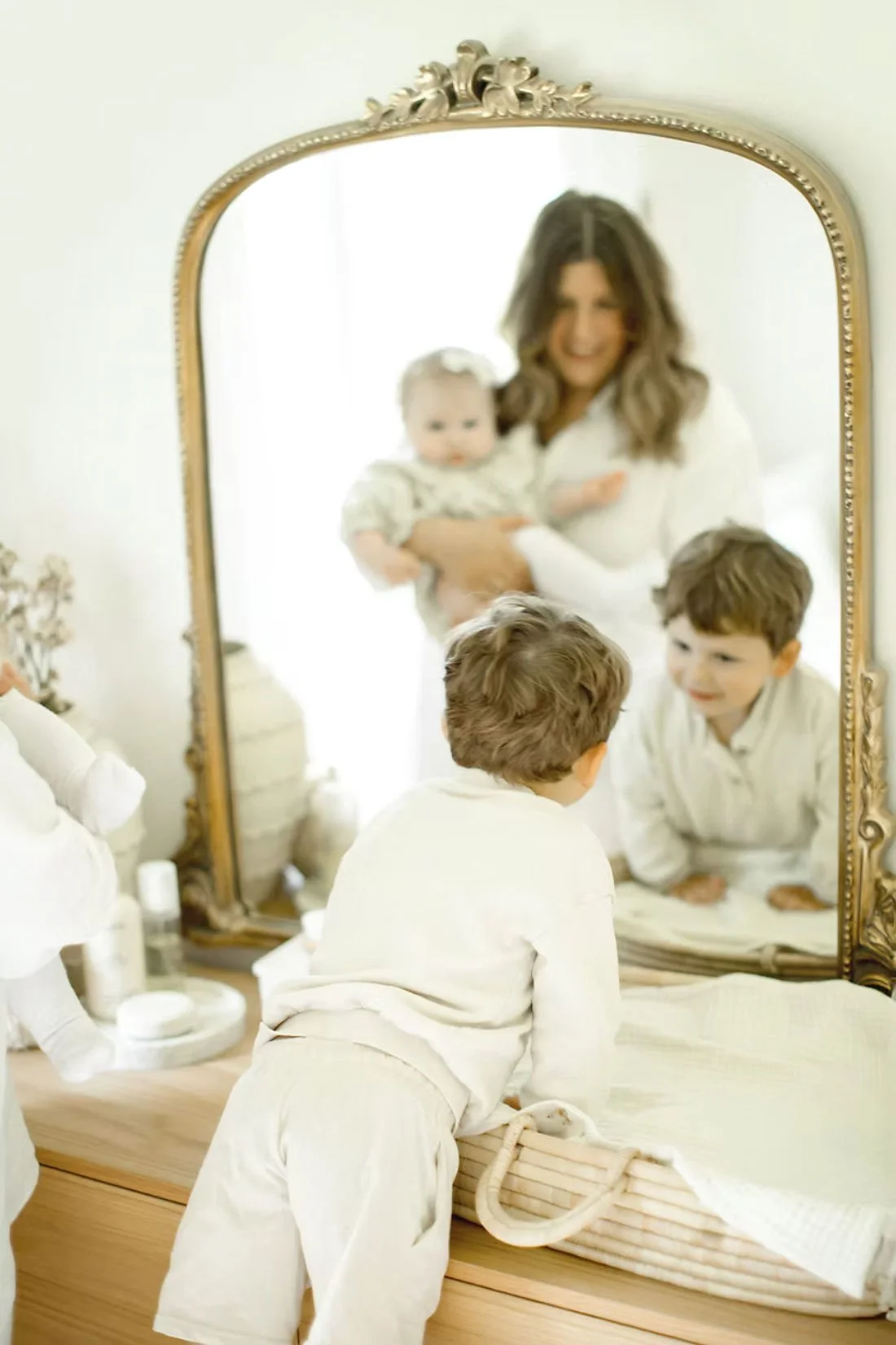 A woman and two children looking at their reflections in a mirror, with the woman holding a baby, all dressed in white pajamas in a bright room.