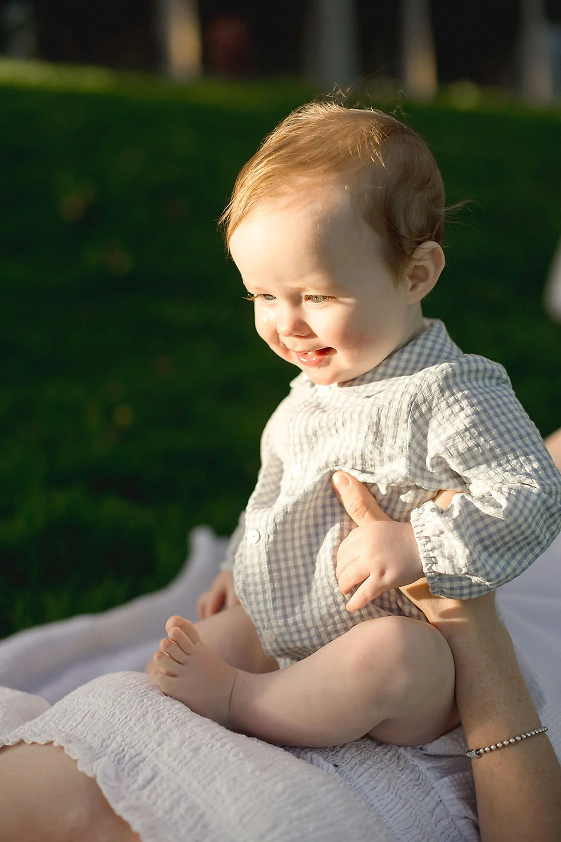 A happy young child with light brown hair, sitting outdoors on a blanket, smiling and looking to the side in the sunlight.