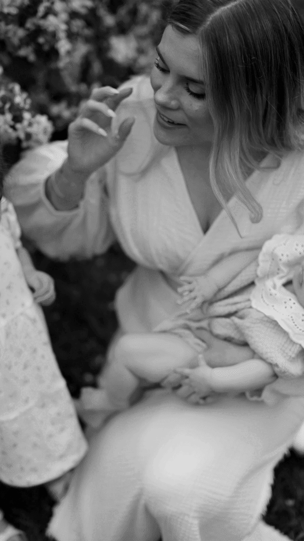 A woman with long hair smiling and holding a baby, with a child nearby, in a garden setting.