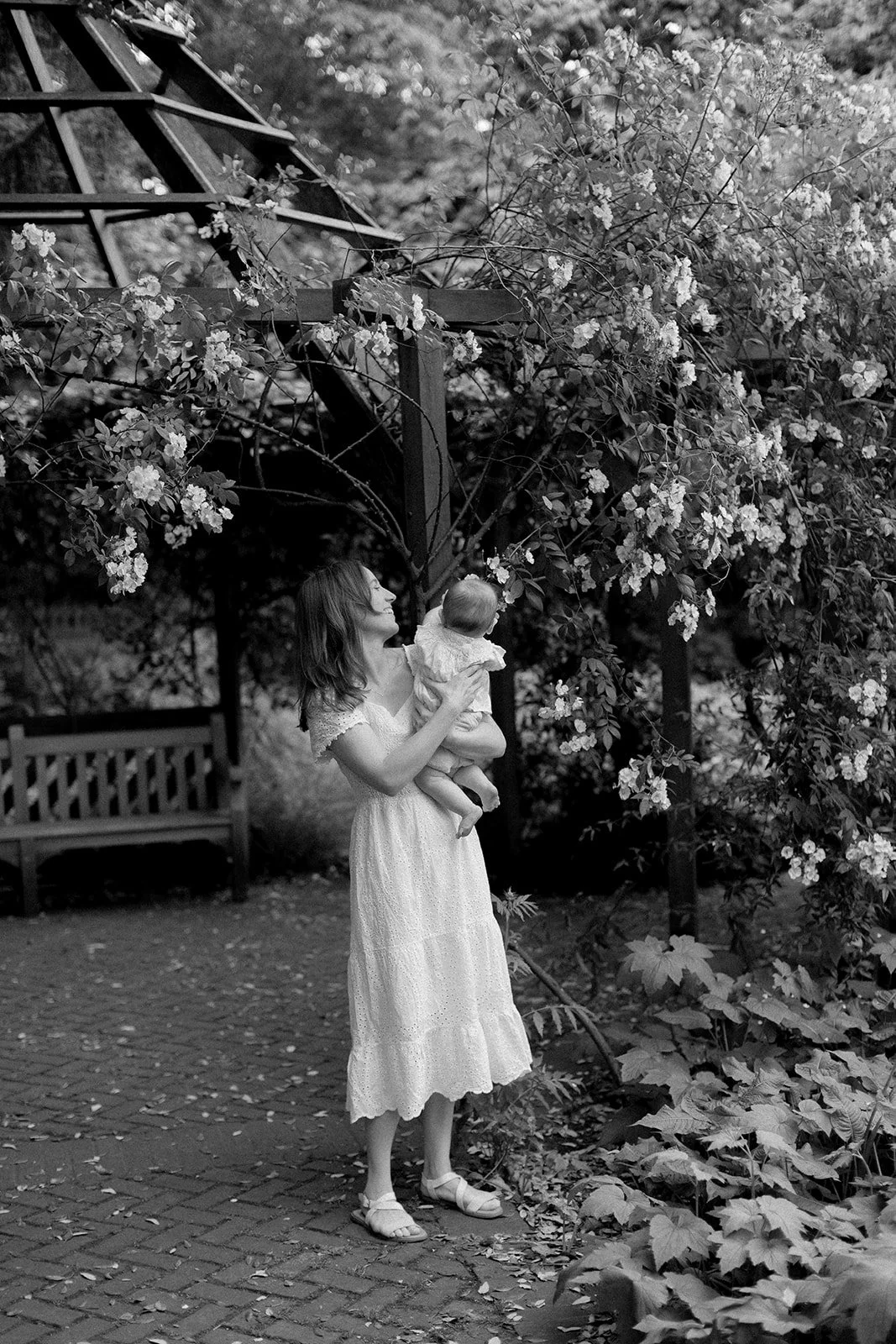 A woman in a white dress holding a baby outdoors near flowering bushes and trees, with a wooden bench in the background in a garden or park setting.