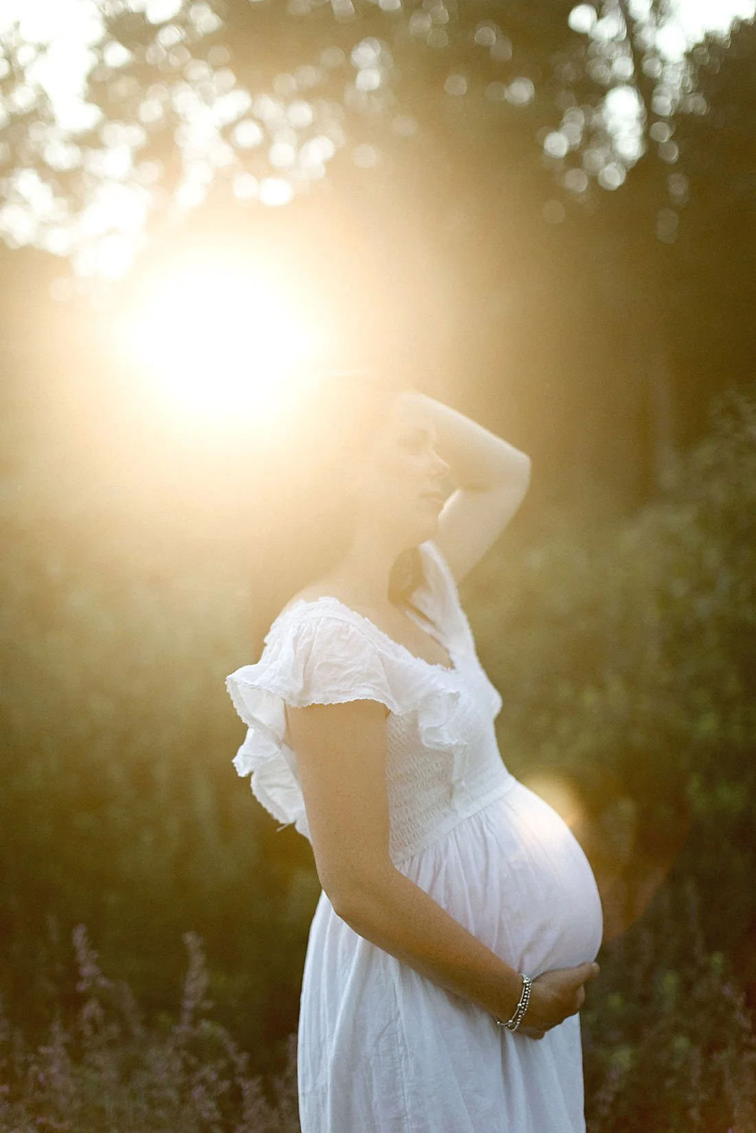 A pregnant woman wearing a white dress outdoors at sunset, with sunlight shining behind her and trees in the background.