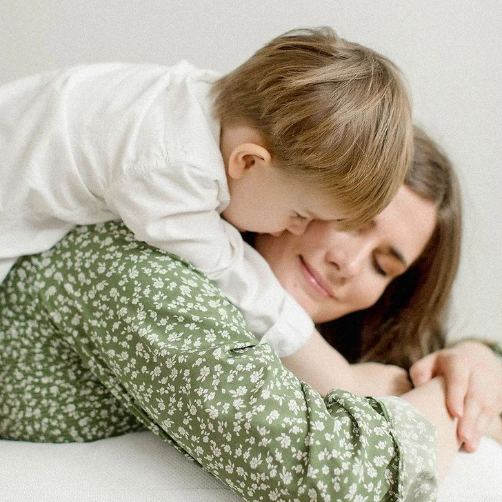 A child hugging and playing with their mother, both smiling, lying on a bed.