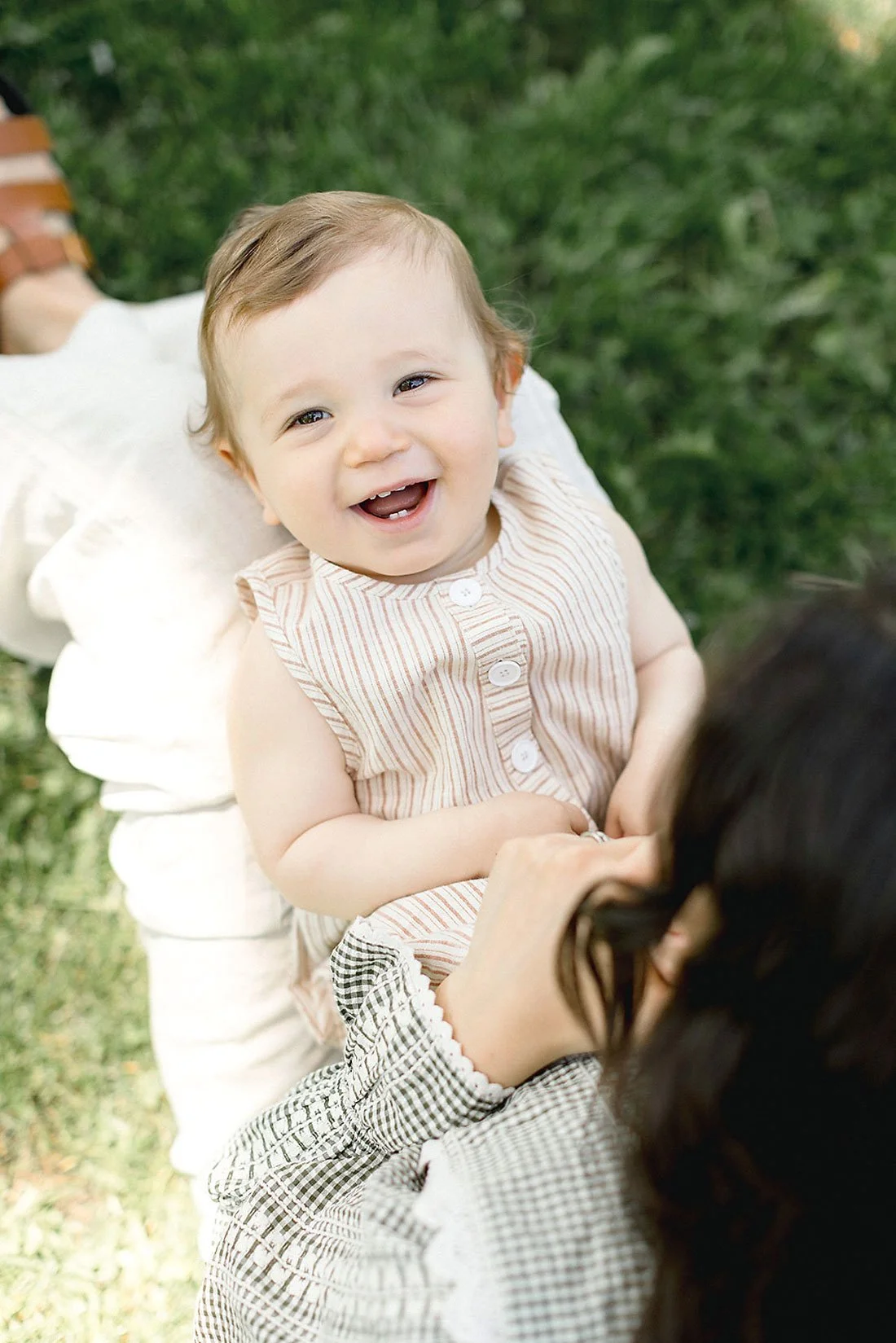 A young child with short light brown hair and a striped sleeveless dress, smiling and looking up at an adult with dark hair and glasses, outdoors on grass.