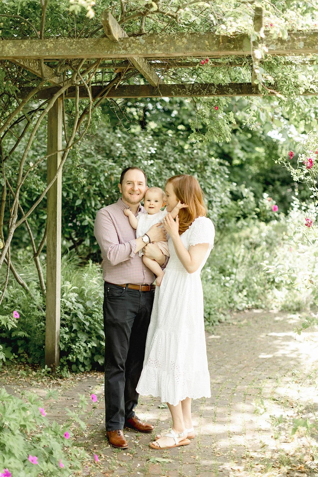 A family of three, a man, woman, and baby, standing outdoors in a lush garden with greenery and pink flowers. The man is holding the baby, and the woman is leaning in, appearing affectionate. They are smiling and dressed in casual summer clothing.