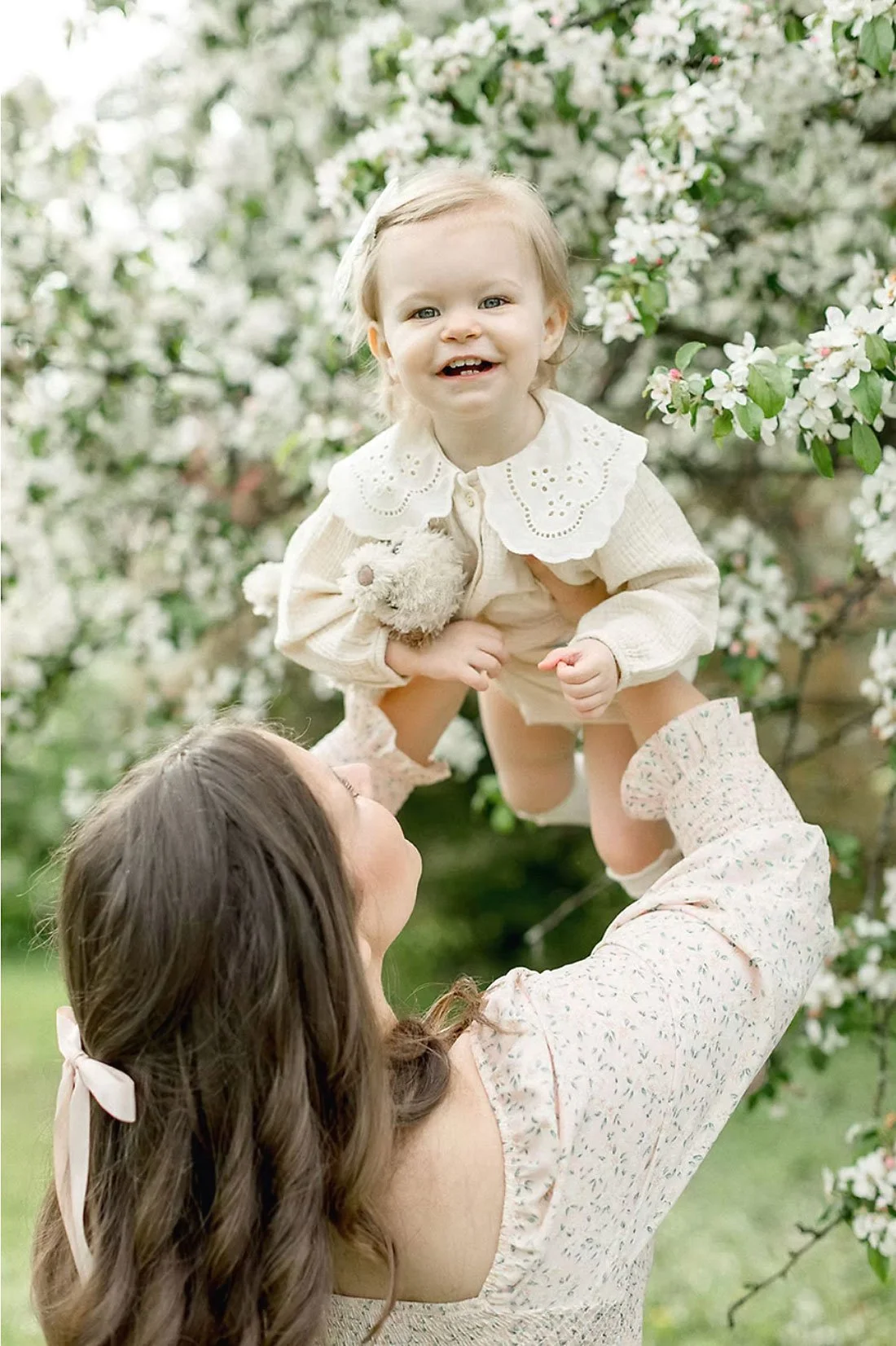 A woman lifting a smiling toddler girl in the air outdoors among flowering trees.