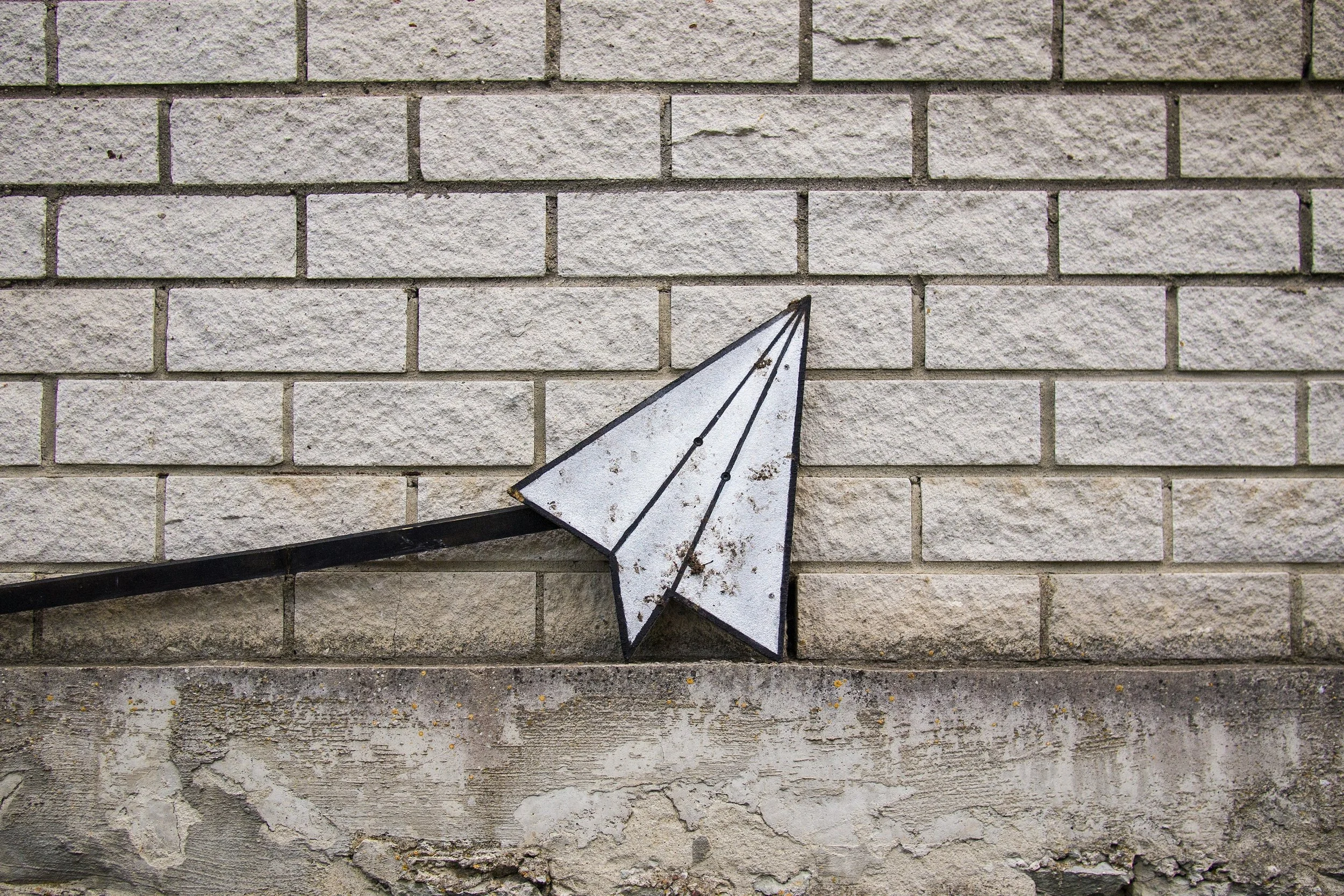 A metal sign in the form of a paper plane in front of a grey brick wall.