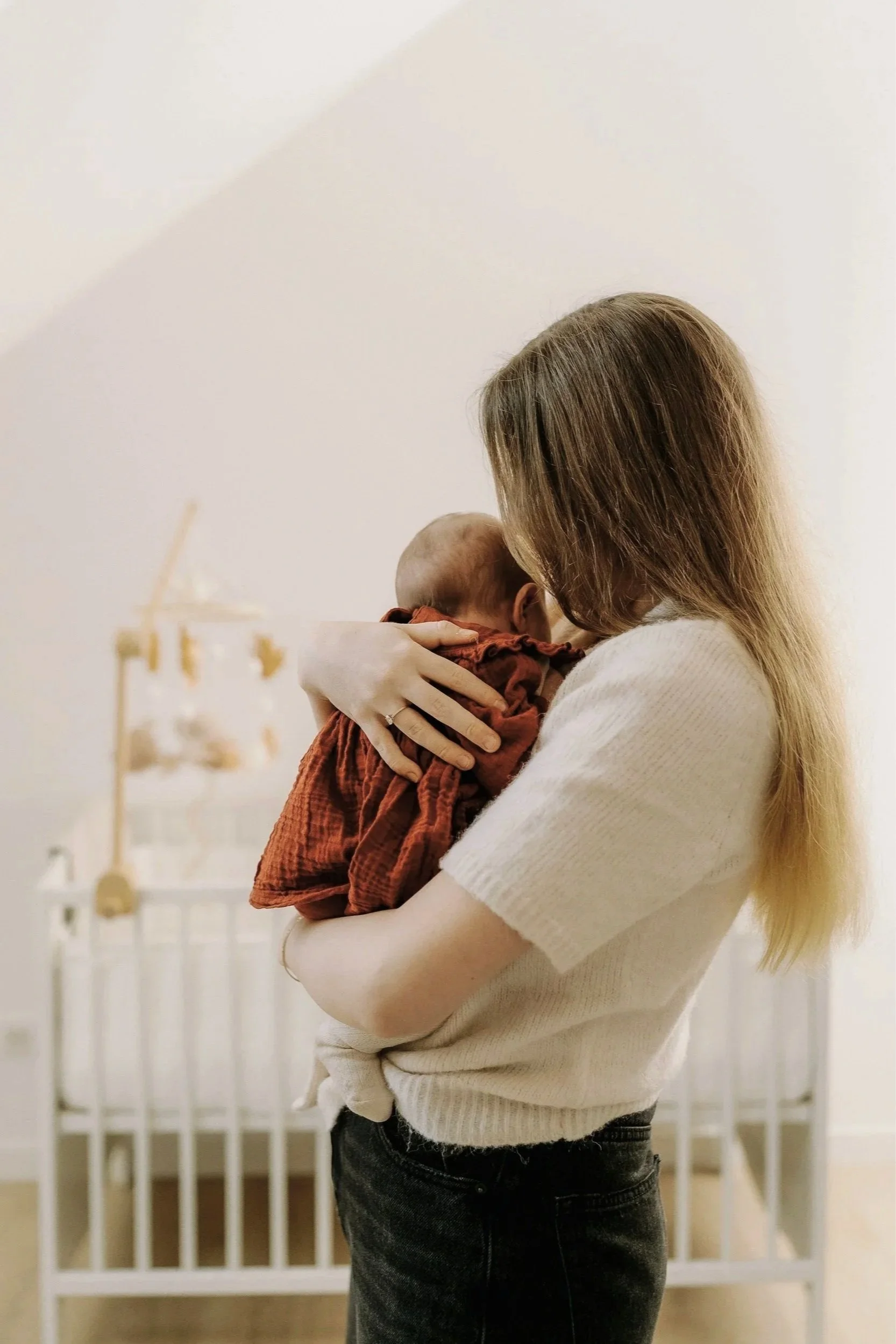 A woman with long blonde hair wearing a cream sweater and black jeans holding a small child dressed in a rust-colored, braided outfit in front of a white crib and a decorative gold and white mobile hanging from the ceiling.