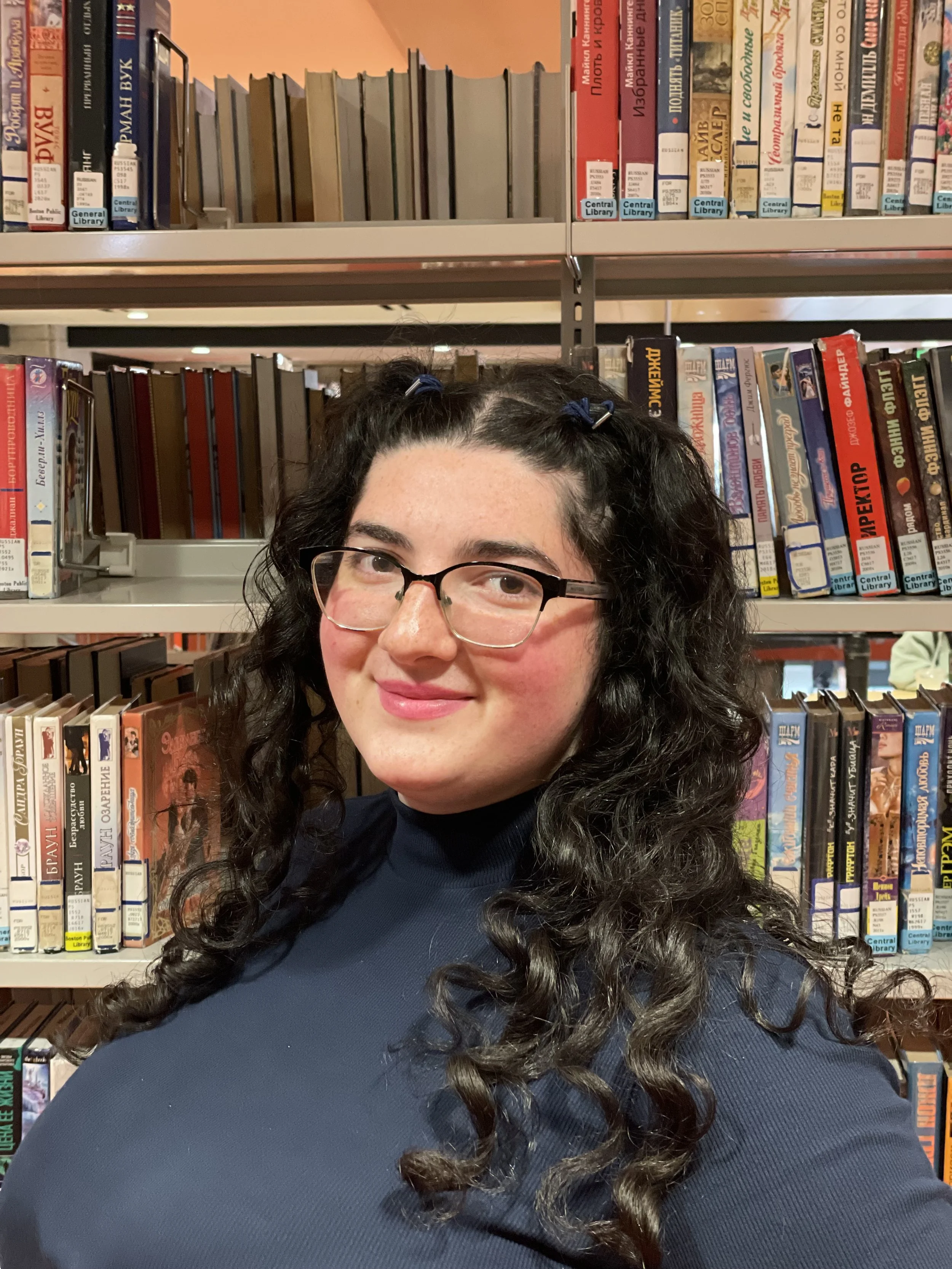 A woman with glasses and curly dark hair smiling in a library with shelves of books behind her.
