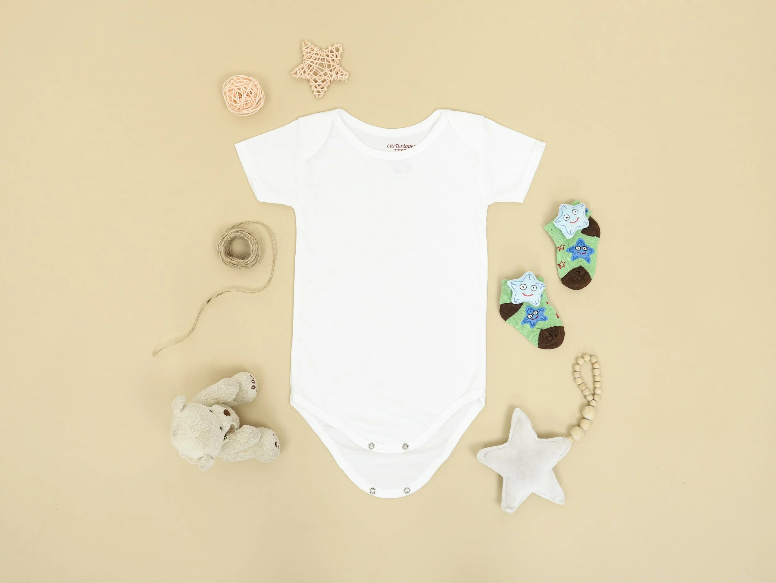 Flat lay of baby items on a beige background including a white baby onesie, plush toys, socks with star faces, a star-shaped pillow, a stuffed bear, yarn balls, and a beaded bracelet.