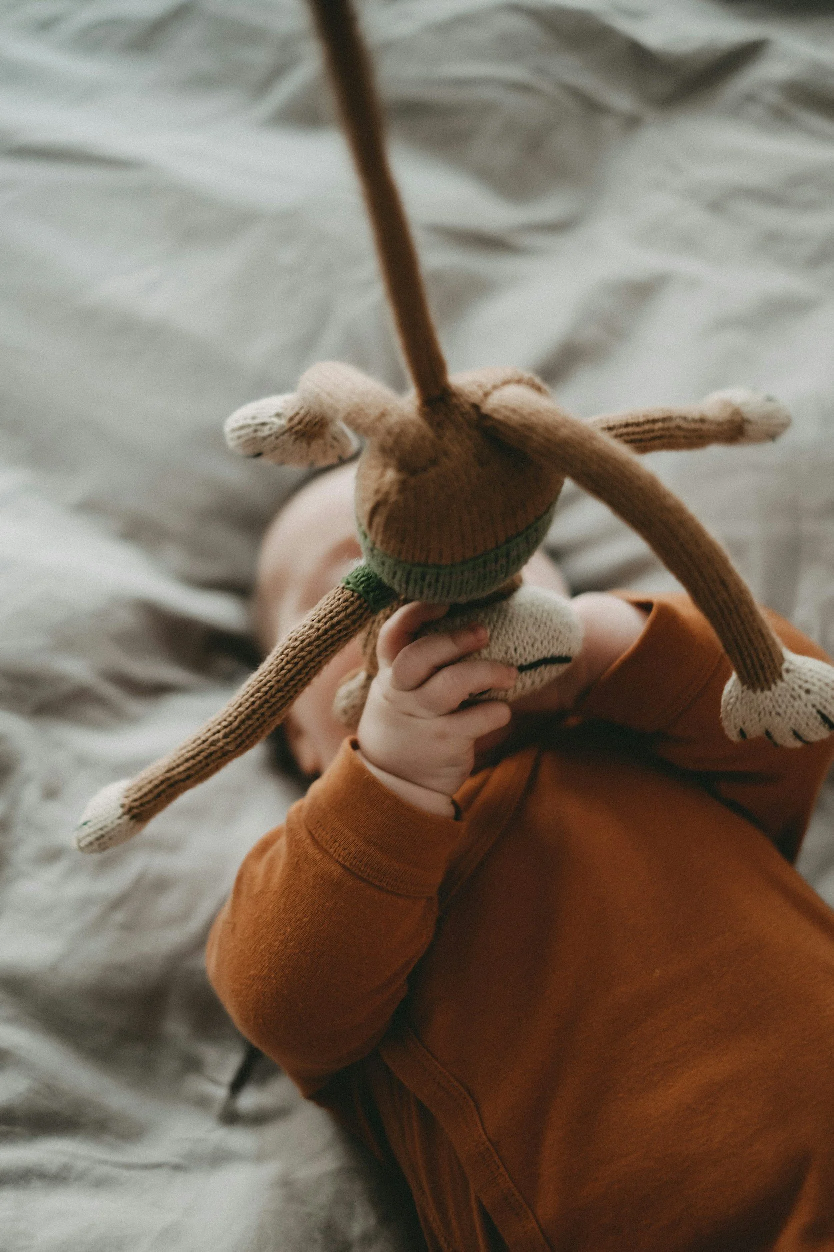 A child dressed in a brown long-sleeve shirt holding a plush dog toy with long limbs, while lying on a soft, light-colored surface.