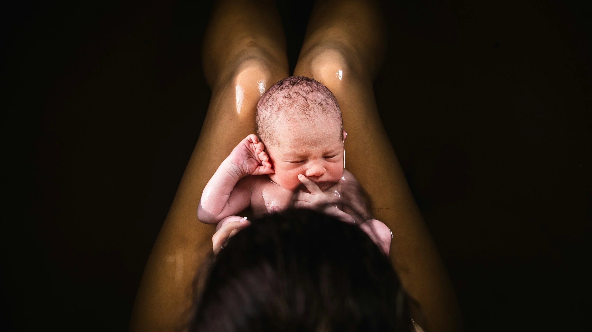 A newborn baby with wet hair lying on someone's legs in a dark environment.