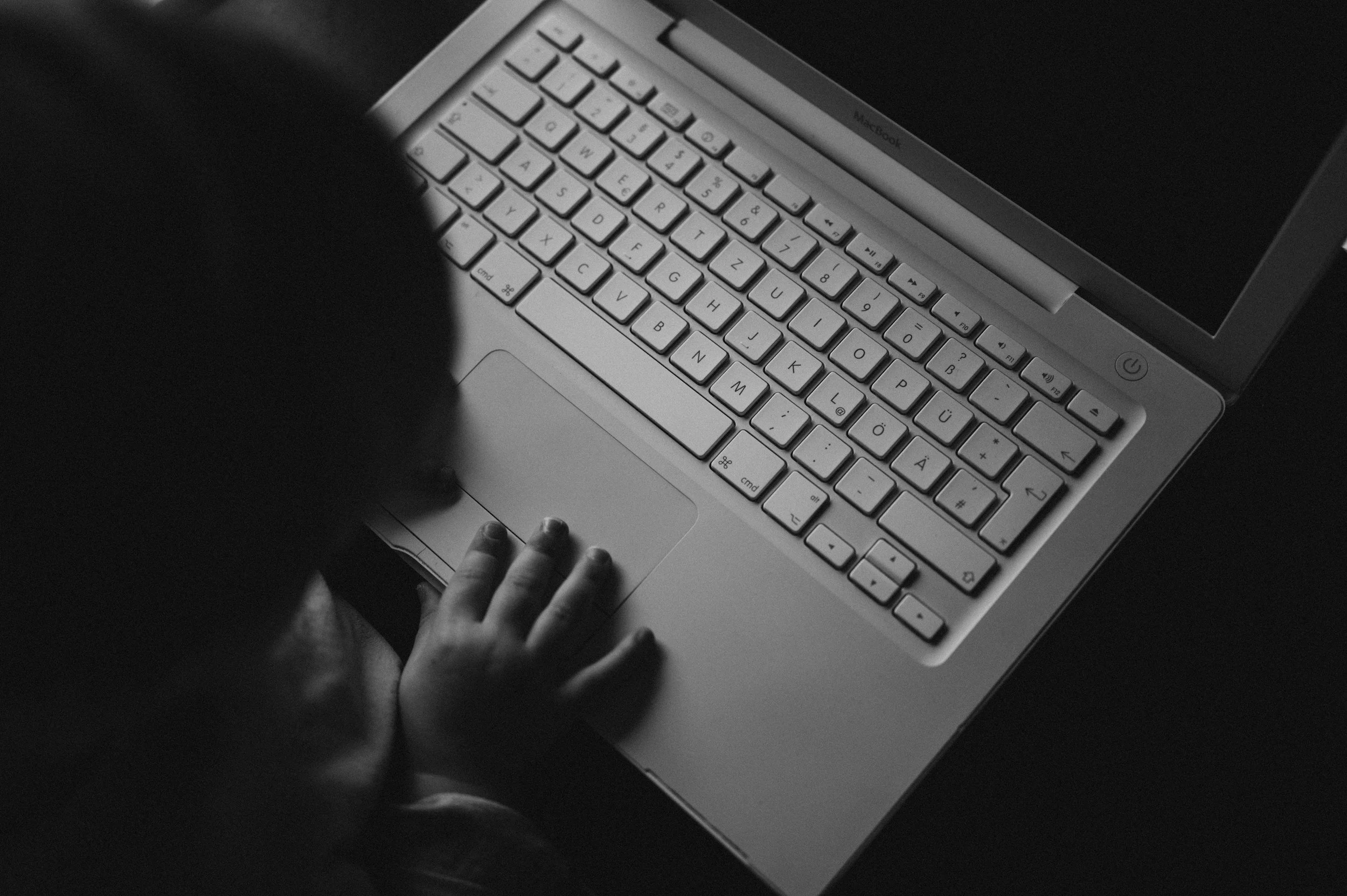 A person’s hand resting on a keyboard of a closed MacBook laptop, which is being held open in a dark environment, with the screen turned off.