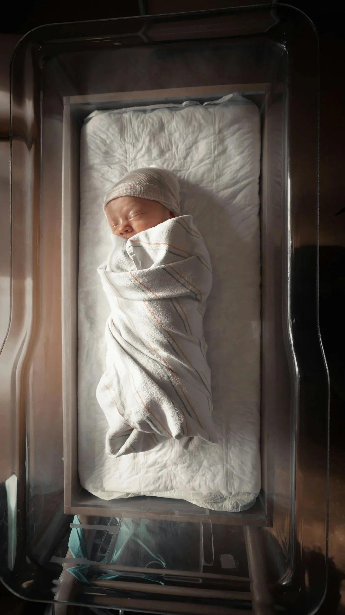 Newborn baby in a hospital bassinet, wrapped in a striped blanket, wearing a beanie, sleeping peacefully.
