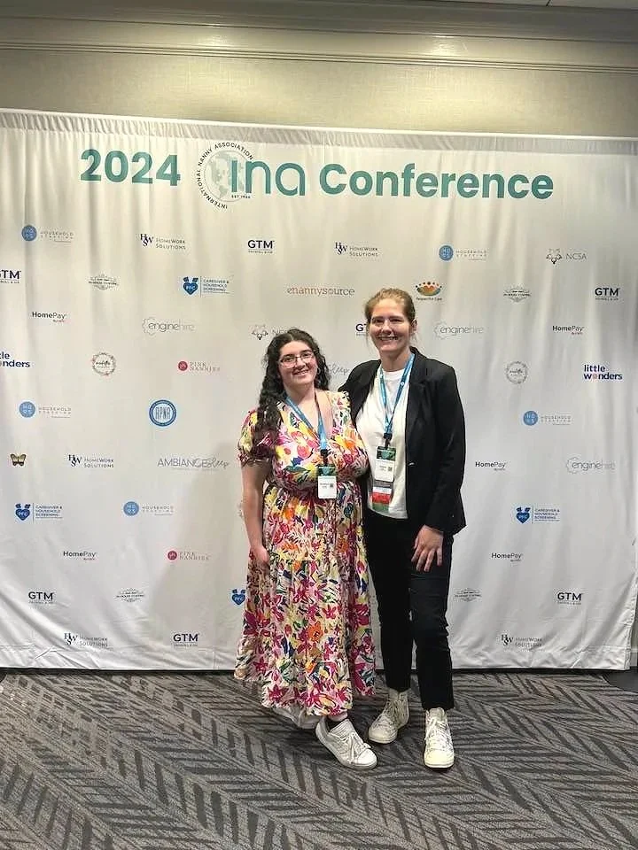 Two women standing together at the 2024 INA Conference, smiling for the photo, with a conference backdrop featuring various sponsor logos in the background.