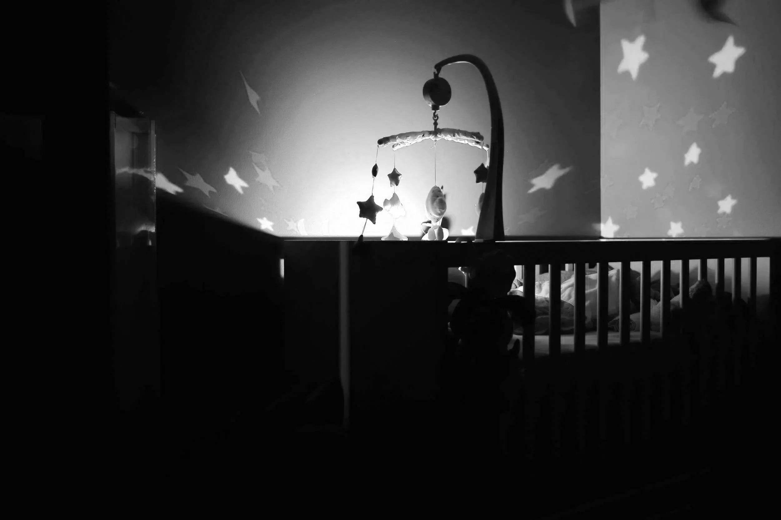 A black and white photo of a baby crib with a mobile hanging above it. The mobile has hanging stars and moon shapes. The background wall has star-shaped patterns projected onto it, creating a gentle and calming nursery scene.