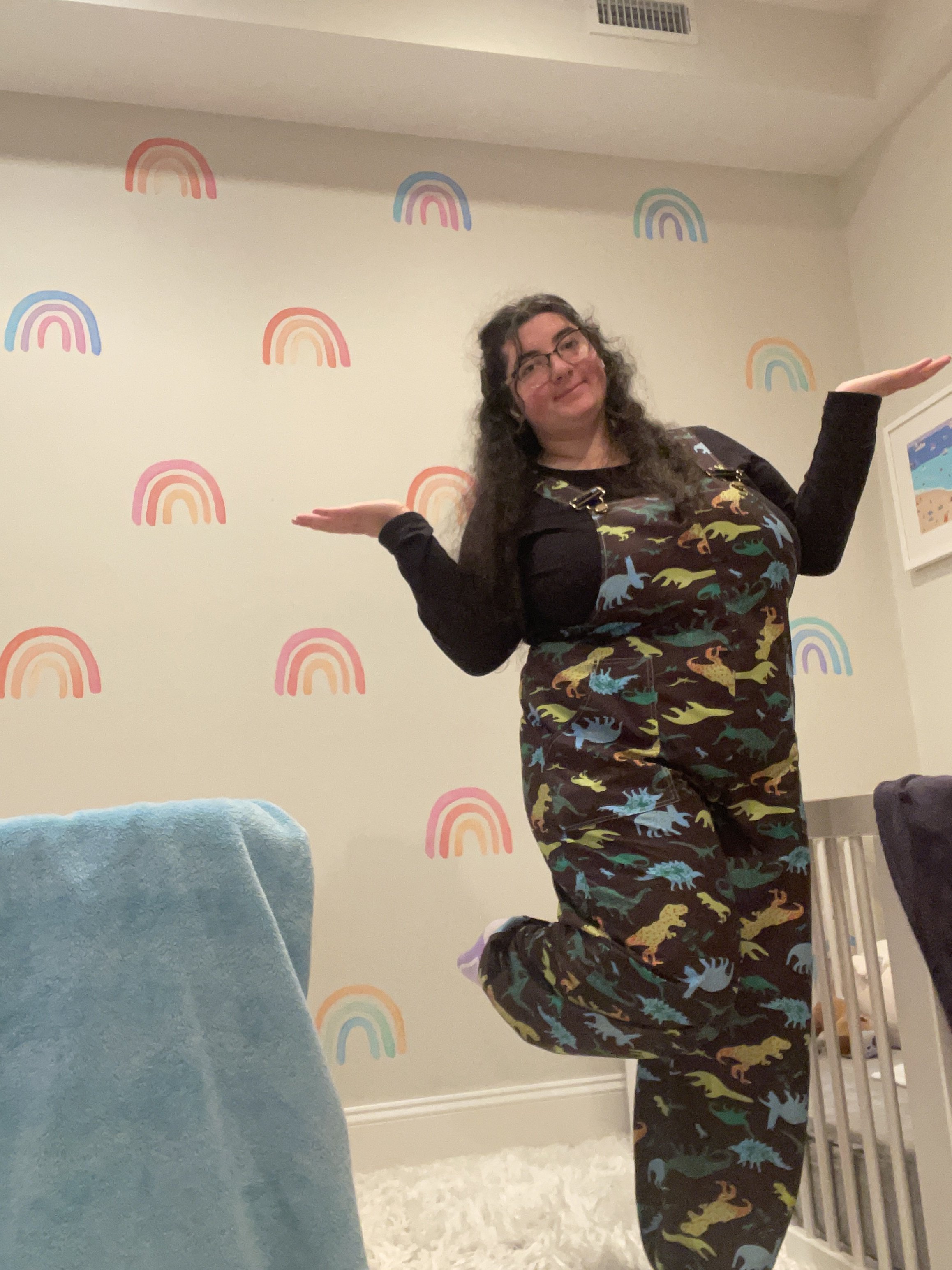 A woman with long wavy hair, glasses, and a dress with dinosaur print standing in a room with a wall decorated with rainbow illustrations, posing with her arms raised and a playful expression.