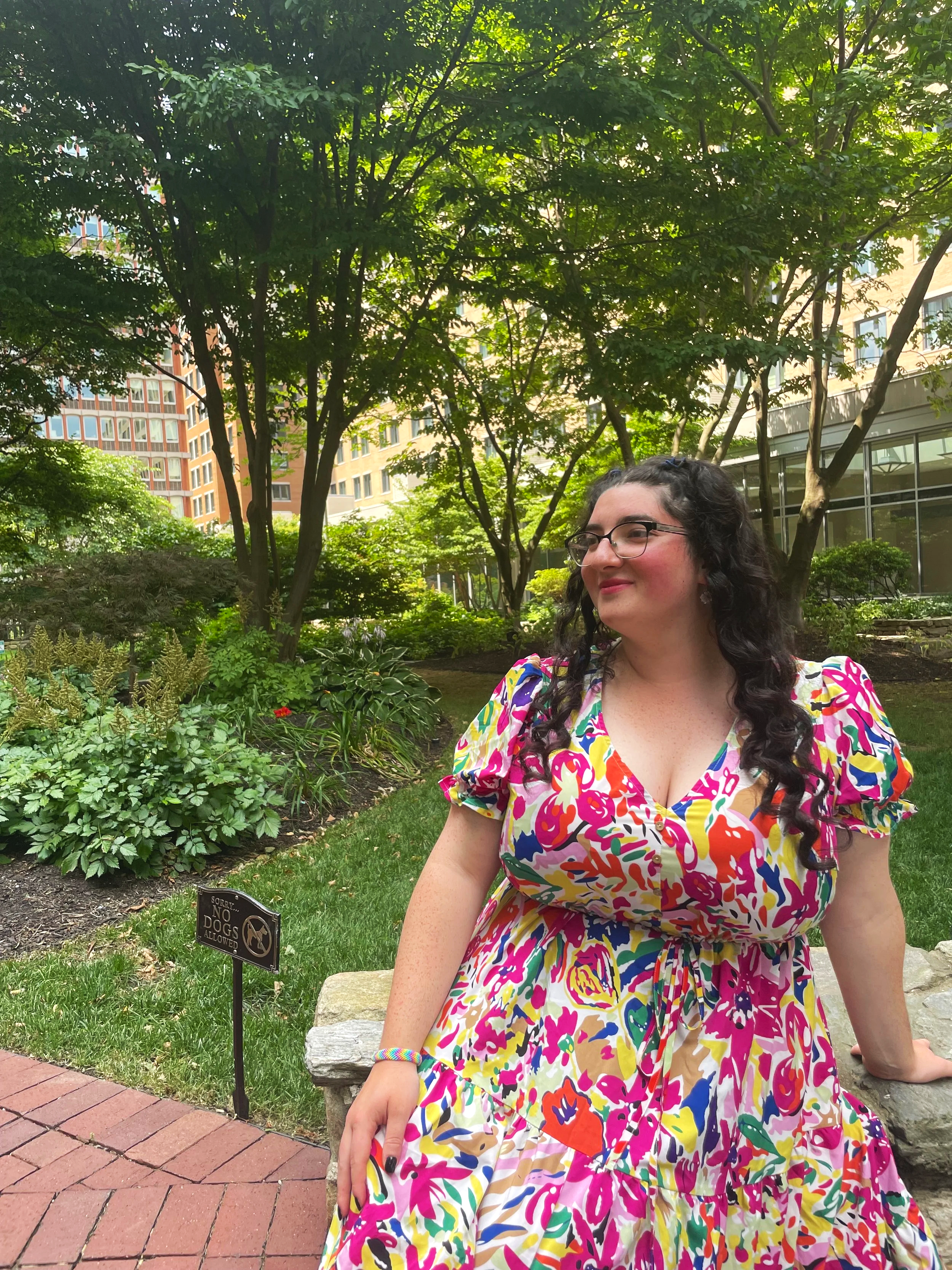 A woman with dark curly hair and glasses wearing a colorful, patterned dress sitting on a stone bench in a green park with trees and buildings in the background. There is a small black sign in the grass that says "Sorry, No Dogs Allowed."