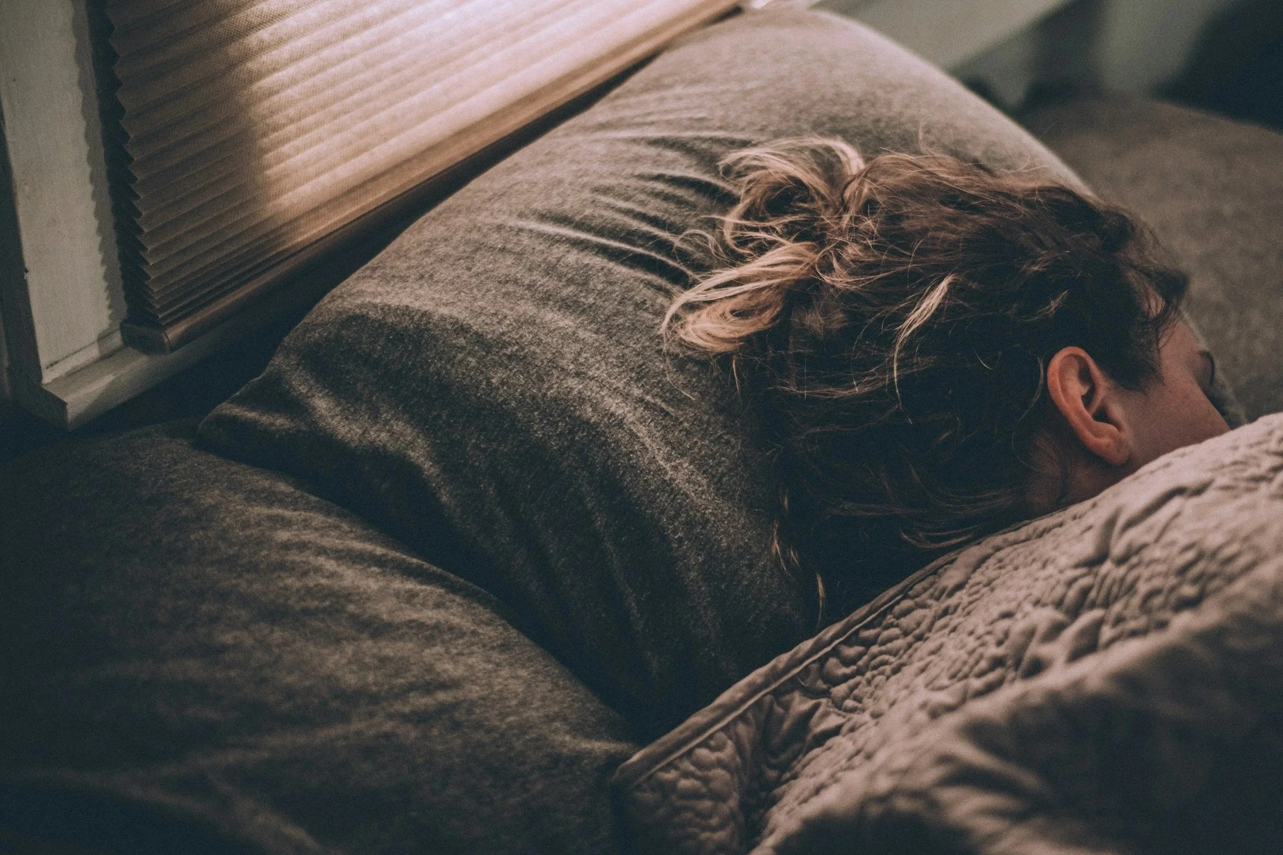 Person sleeping in bed with brown hair, lying on their side on a gray upholstered headboard and pillow, with blinds partially visible above.