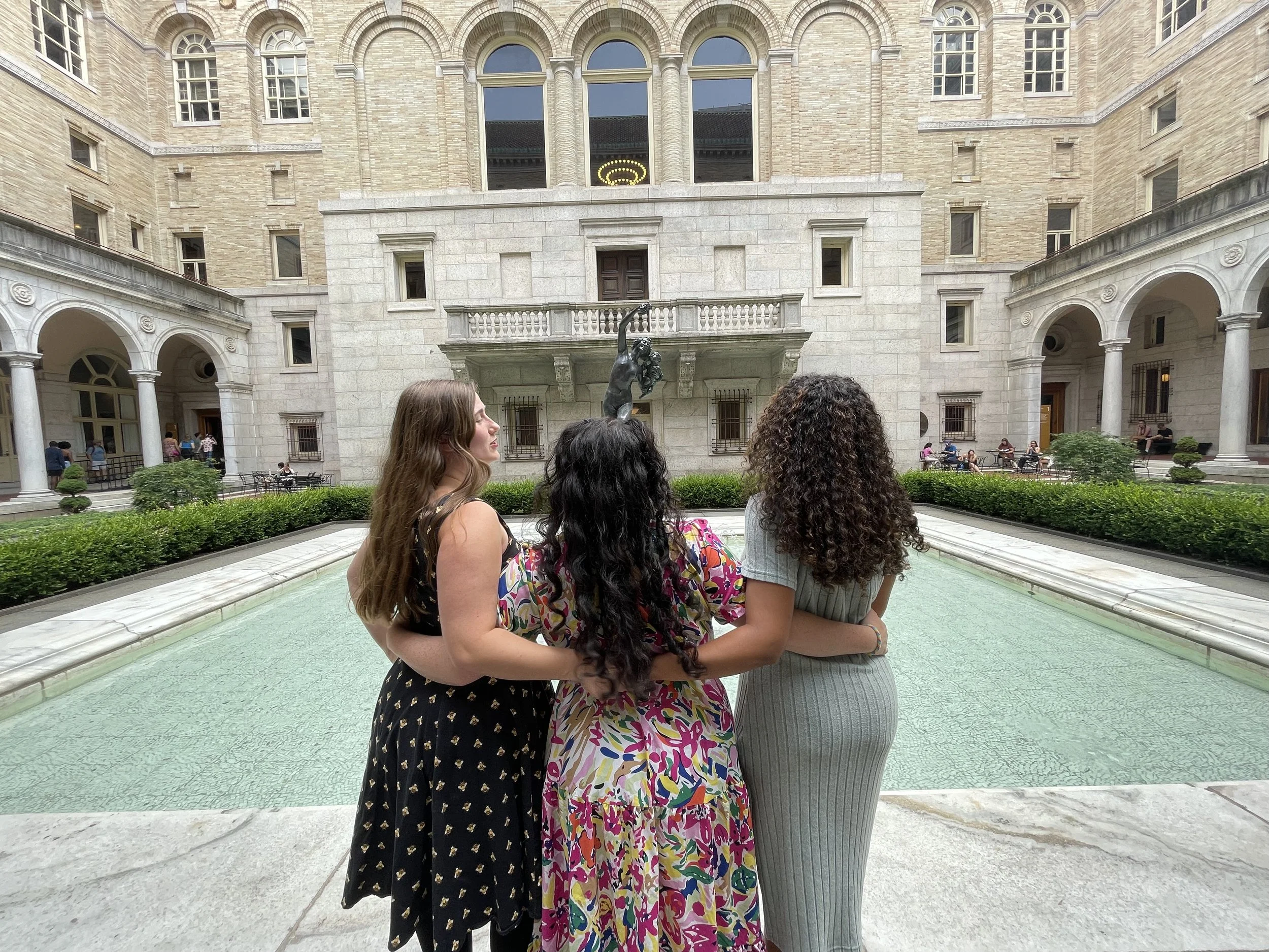 Three women standing by a fountain with a bronze statue of a woman holding a small child in front of a historic brick building with arched windows.