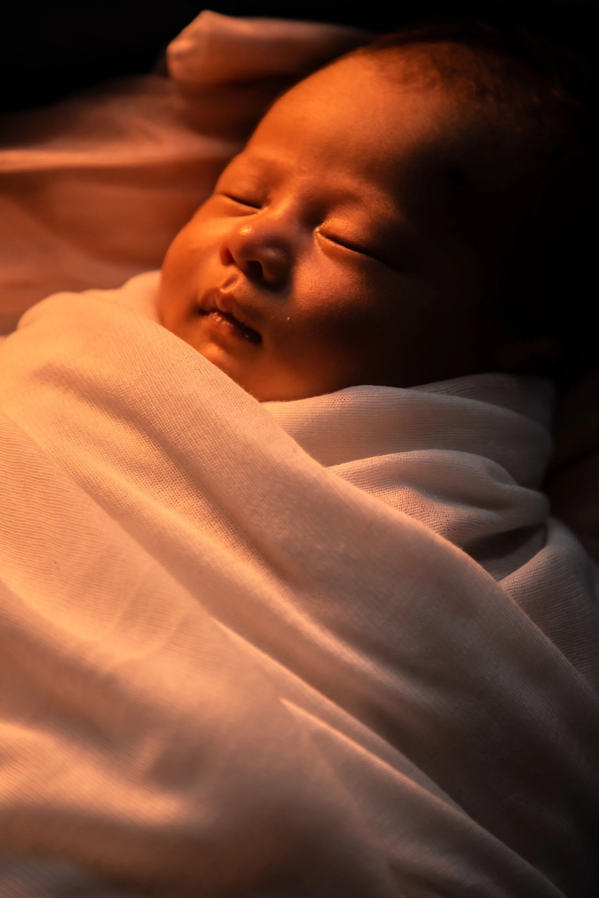 Close-up of a sleeping baby's face, wrapped in a light-colored blanket, with warm, soft lighting.