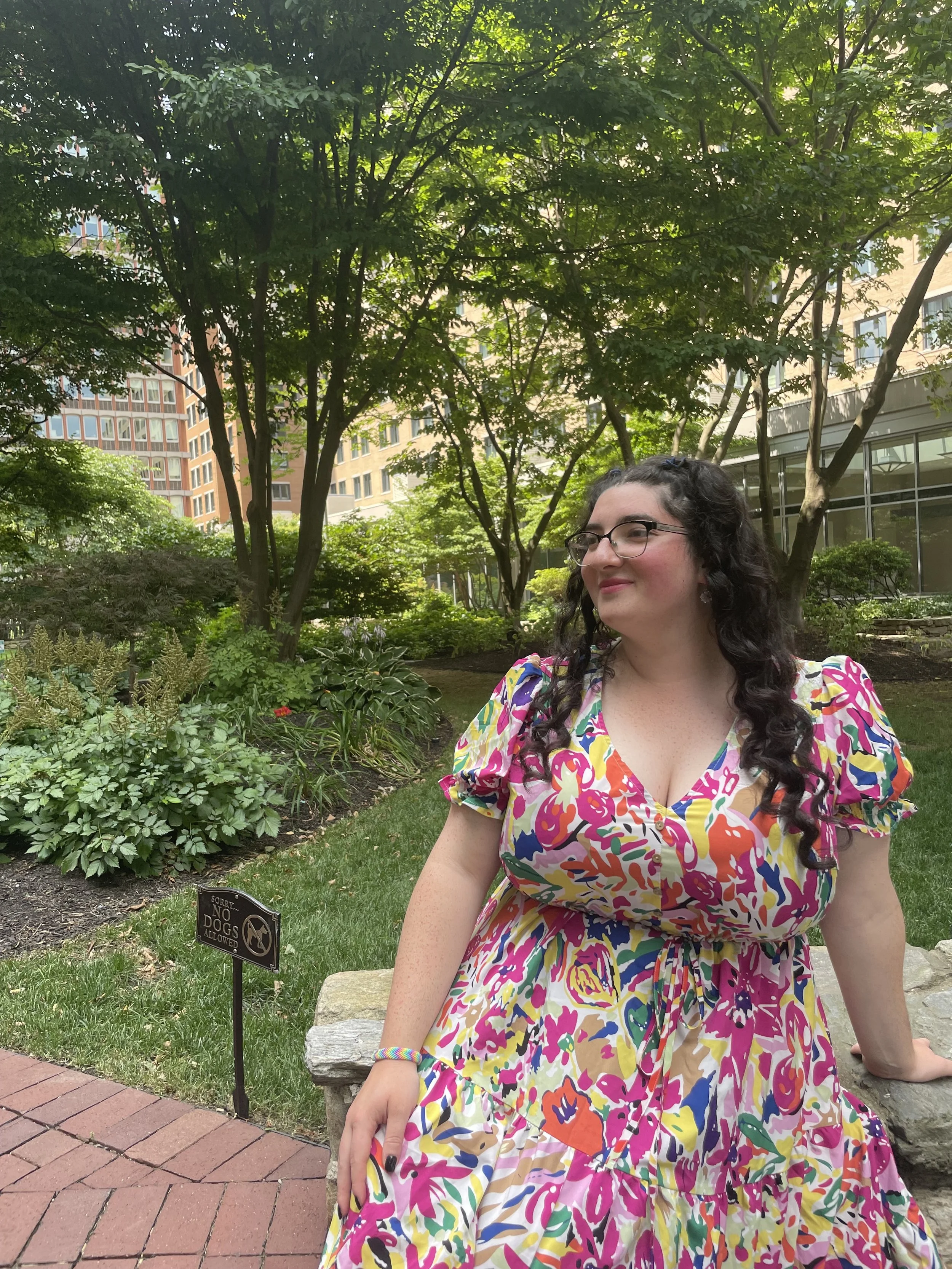 A woman with dark curly hair, glasses, and a colorful dress is sitting on a stone bench in a park, surrounded by trees and greenery, with buildings in the background.