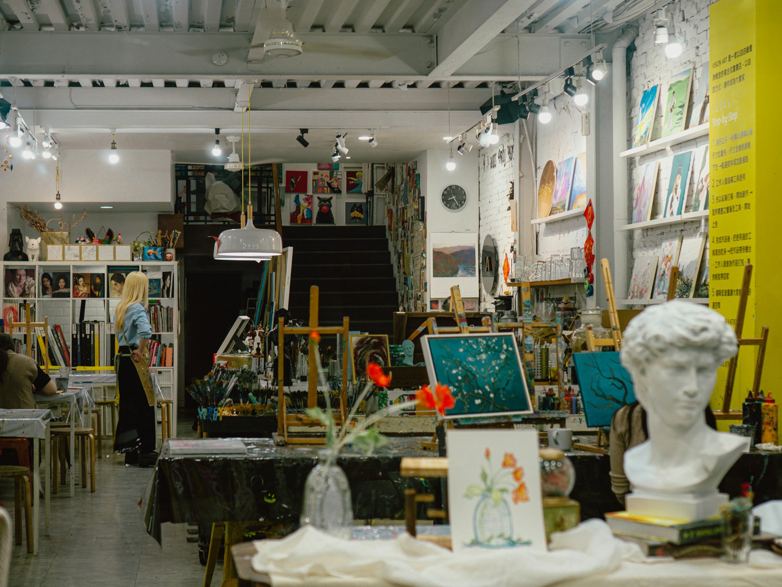Interior of an art gallery or store with paintings, sculptures, and art supplies on display. There are tables with art projects and paintings, and a white brick wall with colorful artwork. Lighting fixtures hang from the ceiling."