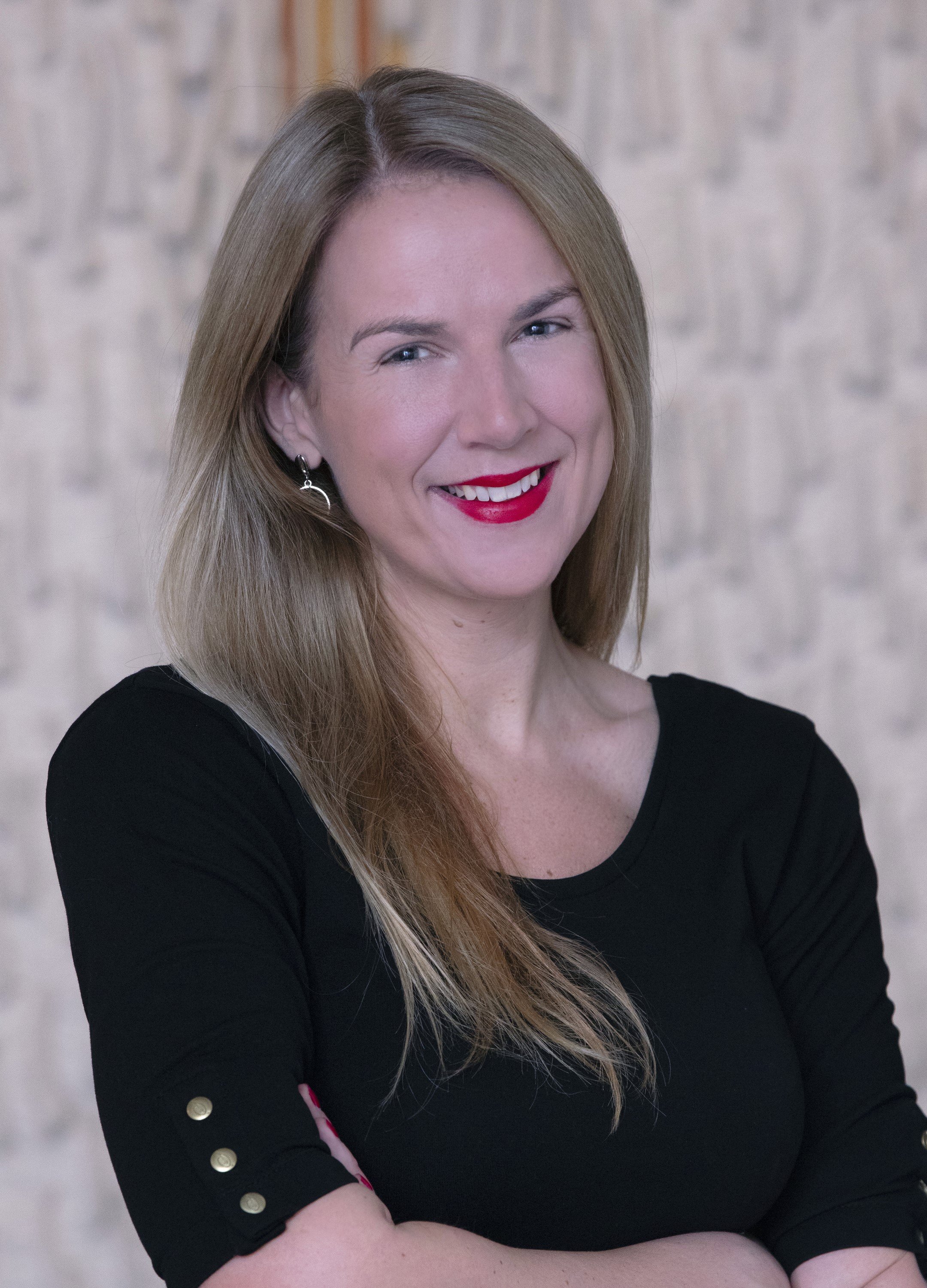 A smiling woman with light brown hair, red lipstick, wearing earrings, and a black top with button details on the sleeves, standing against a light-colored textured background.