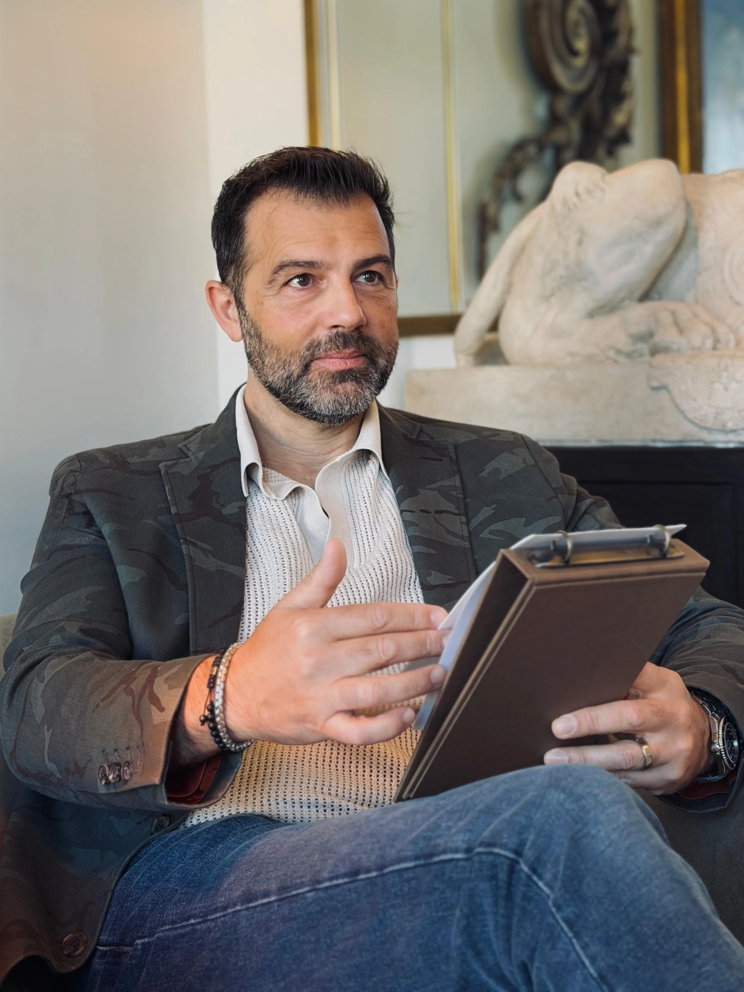 A man with dark hair and a beard sitting on a couch, holding a tablet, wearing a suit jacket, and giving a thumbs-up gesture.