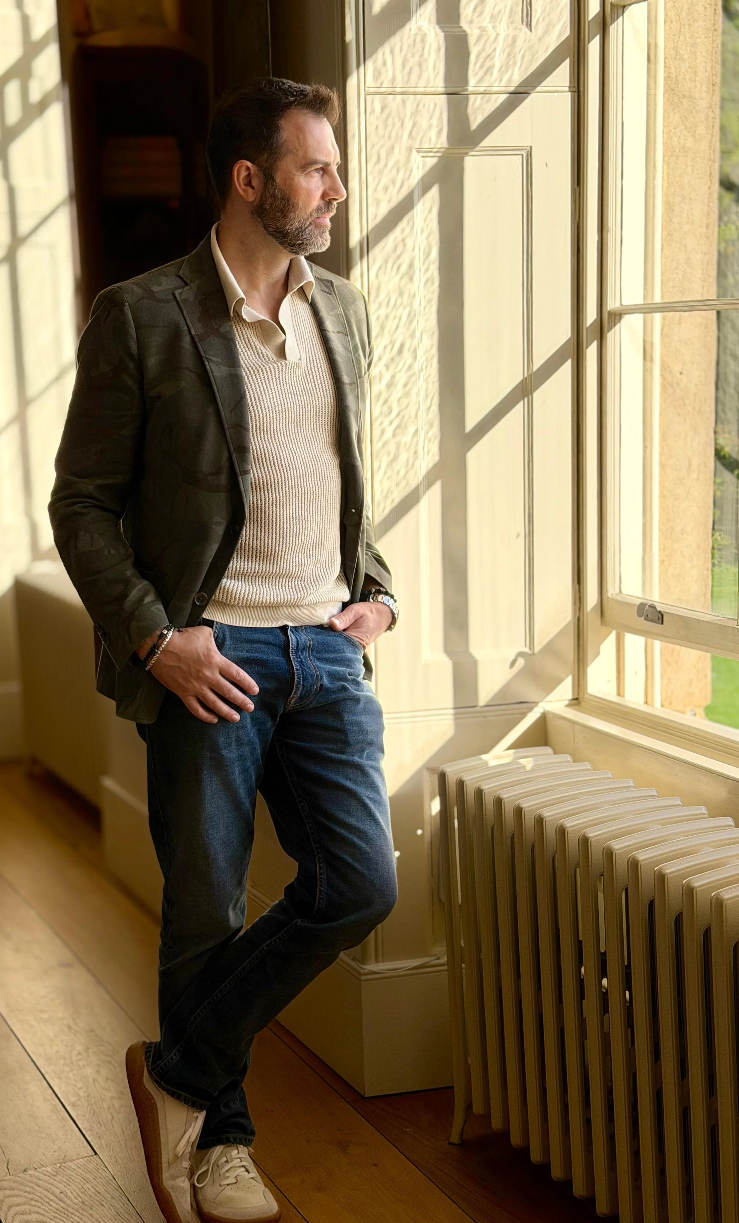 A man with a beard wearing a gray blazer, beige sweater, and jeans stands by a window with sunlight streaming in, looking outside with hands in pockets.