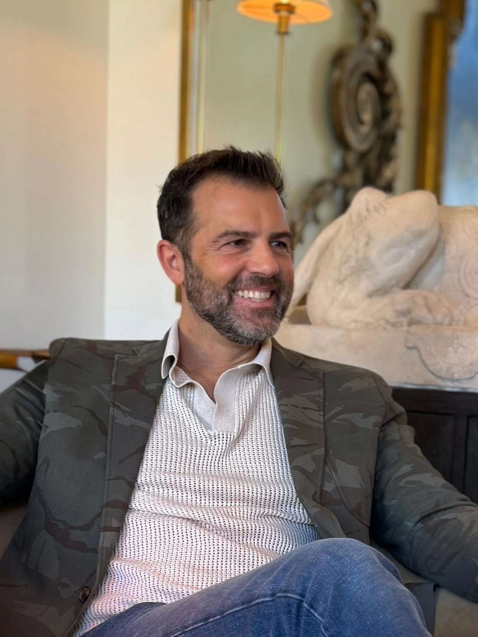 A smiling man with a beard and dark hair, wearing a patterned blazer and white shirt, sitting on a sofa in a room with art and a lamp in the background.