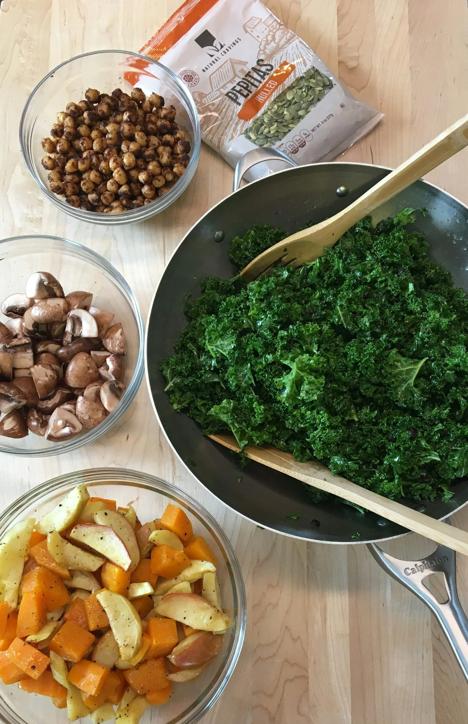 A wooden table with a pan of chopped kale, bowls of sliced mushrooms, roasted chickpeas, and roasted sweet potatoes and apples, along with a bag of pumpkin seeds.