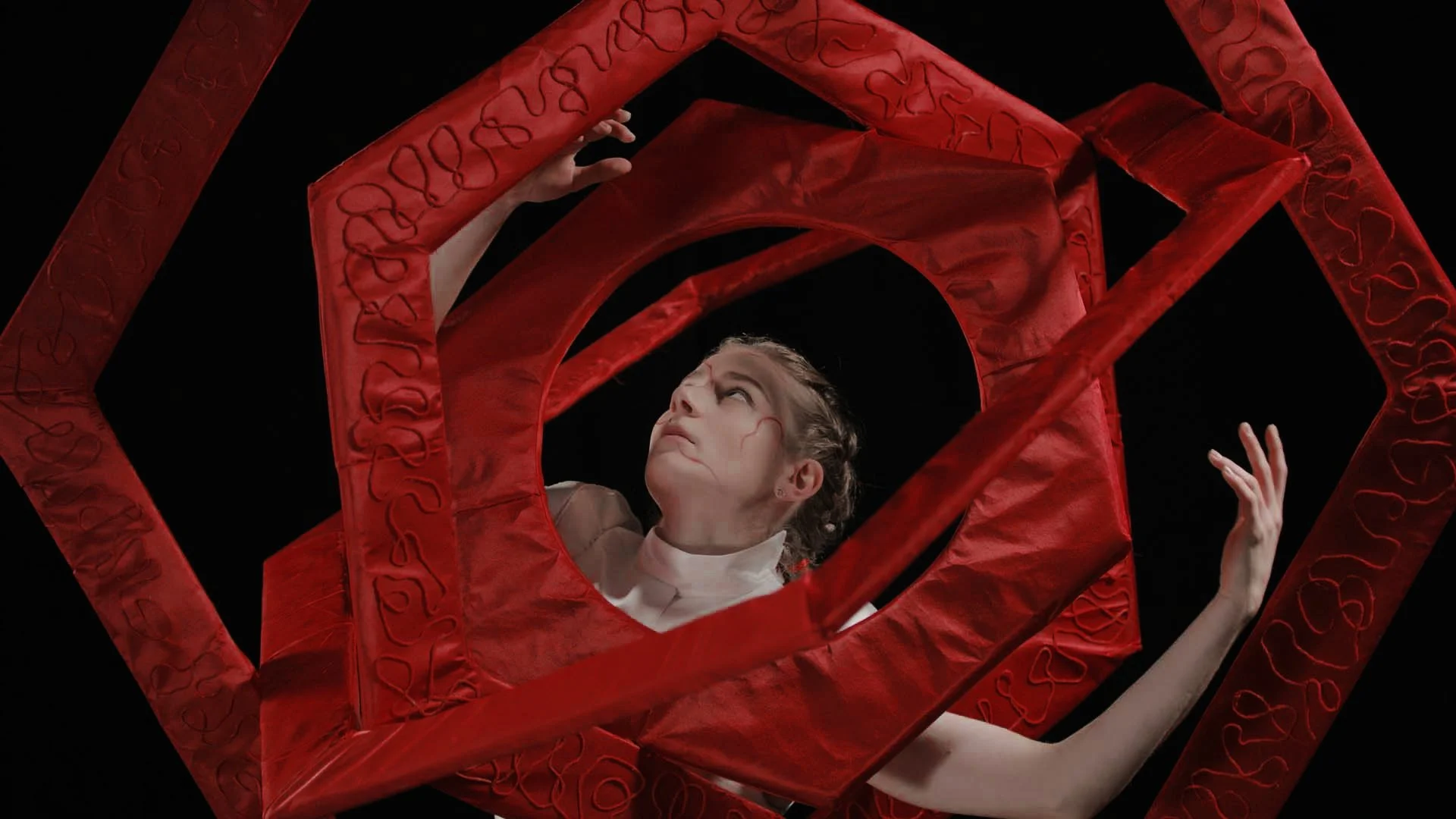 A female dancer with braided hair and wearing a matador costume is surrounded by large red gyroscopic frames on a black background. She is looking upward with her hand delicately raised.