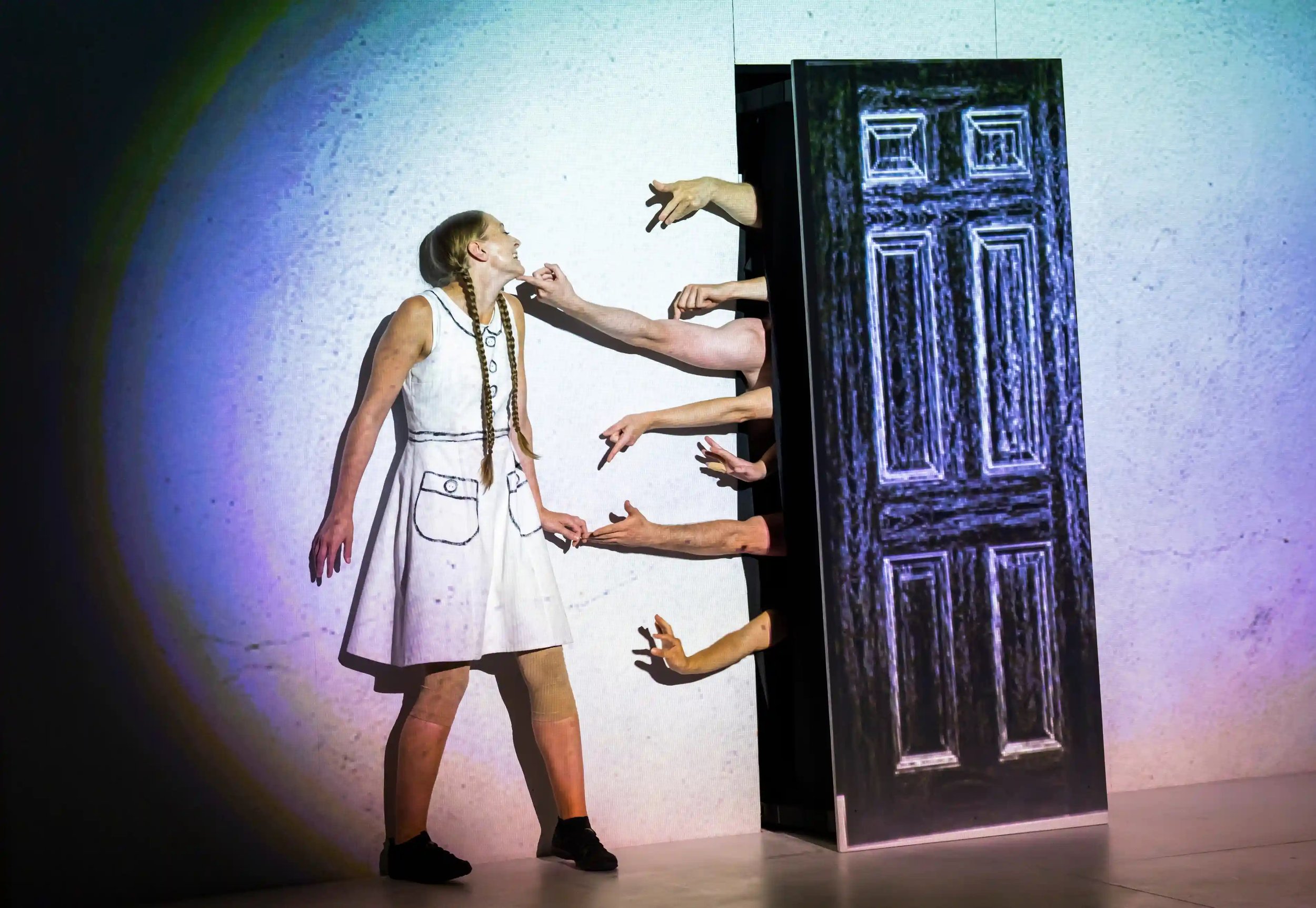 A woman dressed in a white dress with drawn-on pockets standing on stage, smiling and interacting with three people whose hands are reaching out from behind a patterned black-and-white screen or partition.