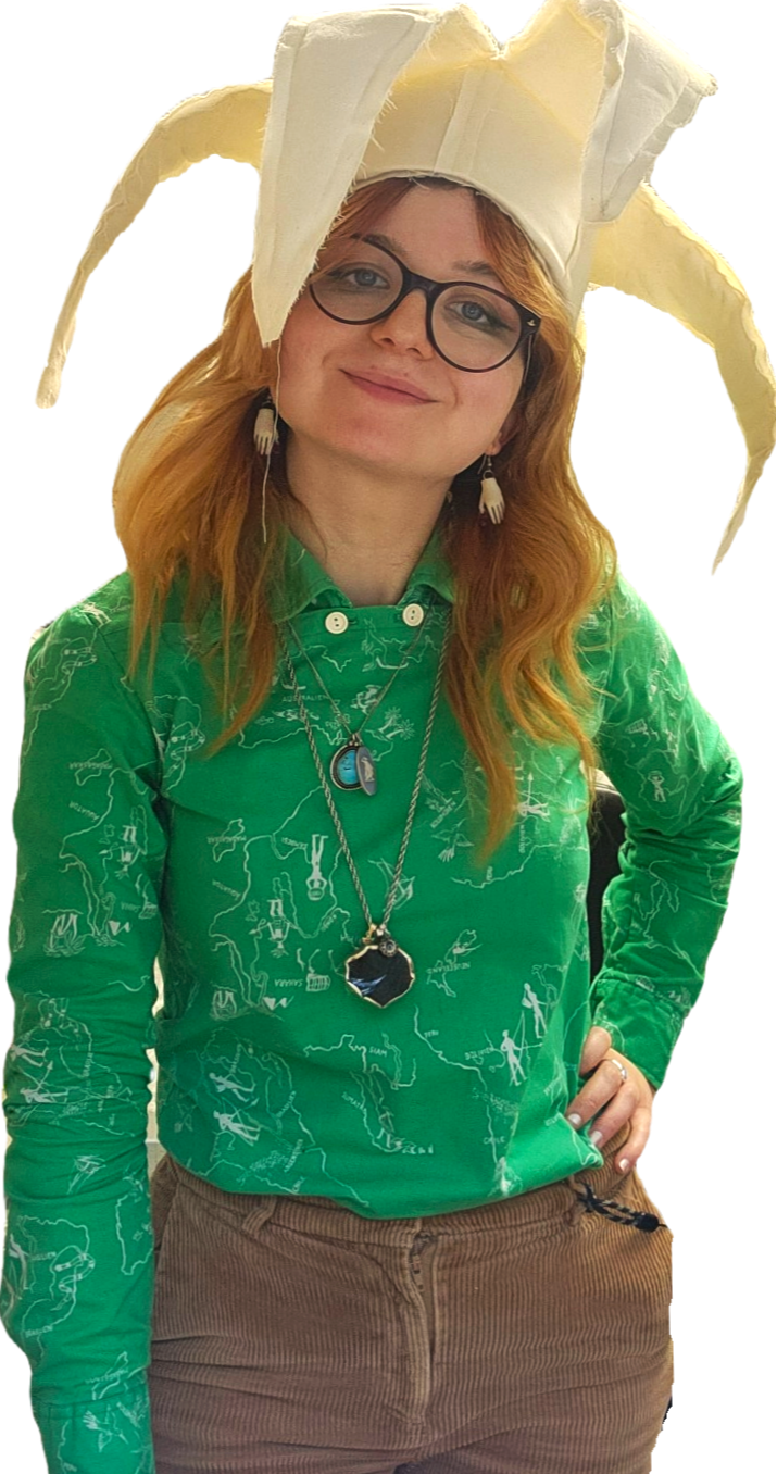 A woman with red hair and glasses wearing a jester hat made of white fabric and a green shirt with white line drawings of maps and figures, accessorized with layered necklaces and earrings shaped like hands.