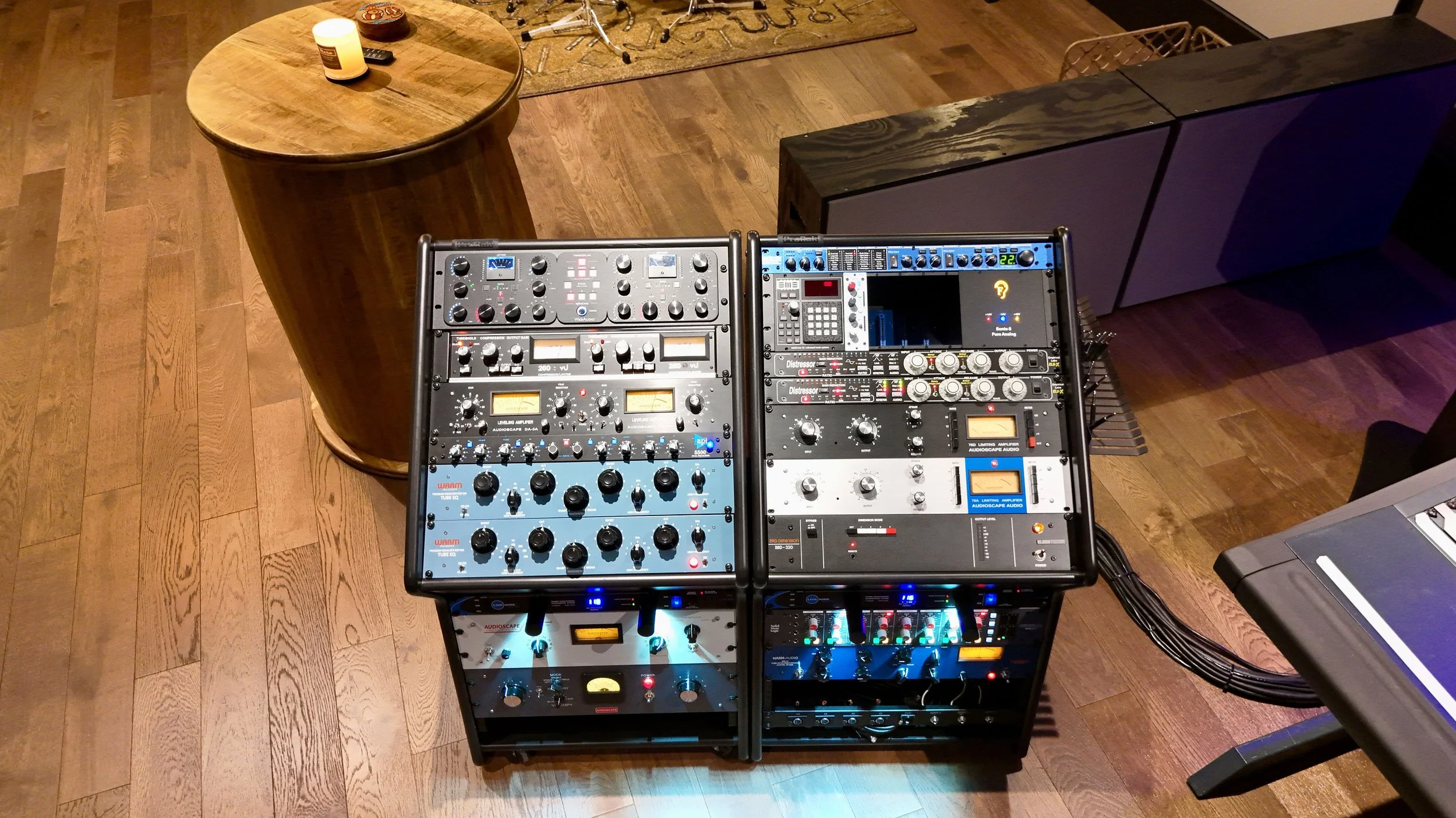 A DJ booth with various audio mixing equipment and controls on a wooden floor, with a small round wooden table and some objects on top nearby.