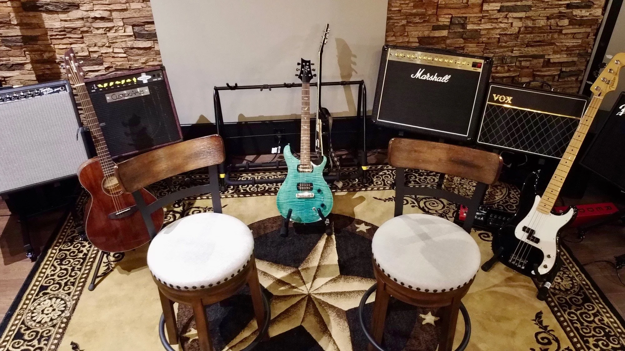 A room with musical instruments including an acoustic guitar, an electric guitar, and an electric bass guitar, along with several amplifiers, chairs, and drums, and a decorative rug and brick wall background.