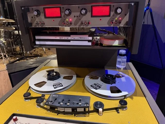 A vintage reel-to-reel tape recorder with two large reels on a yellow surface, a control panel with various knobs and buttons, and an analog level meter. The background includes audio equipment and a water bottle.