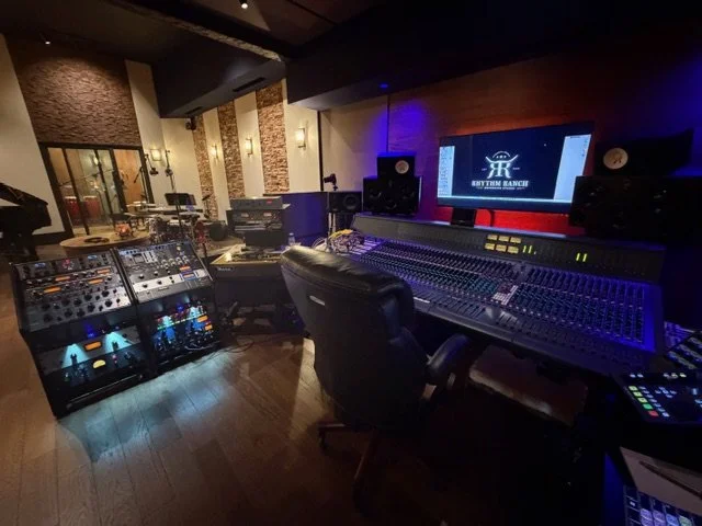 A professional recording studio with mixing console, audio equipment, speakers, and a large screen displaying the Rhythm Ranch logo.