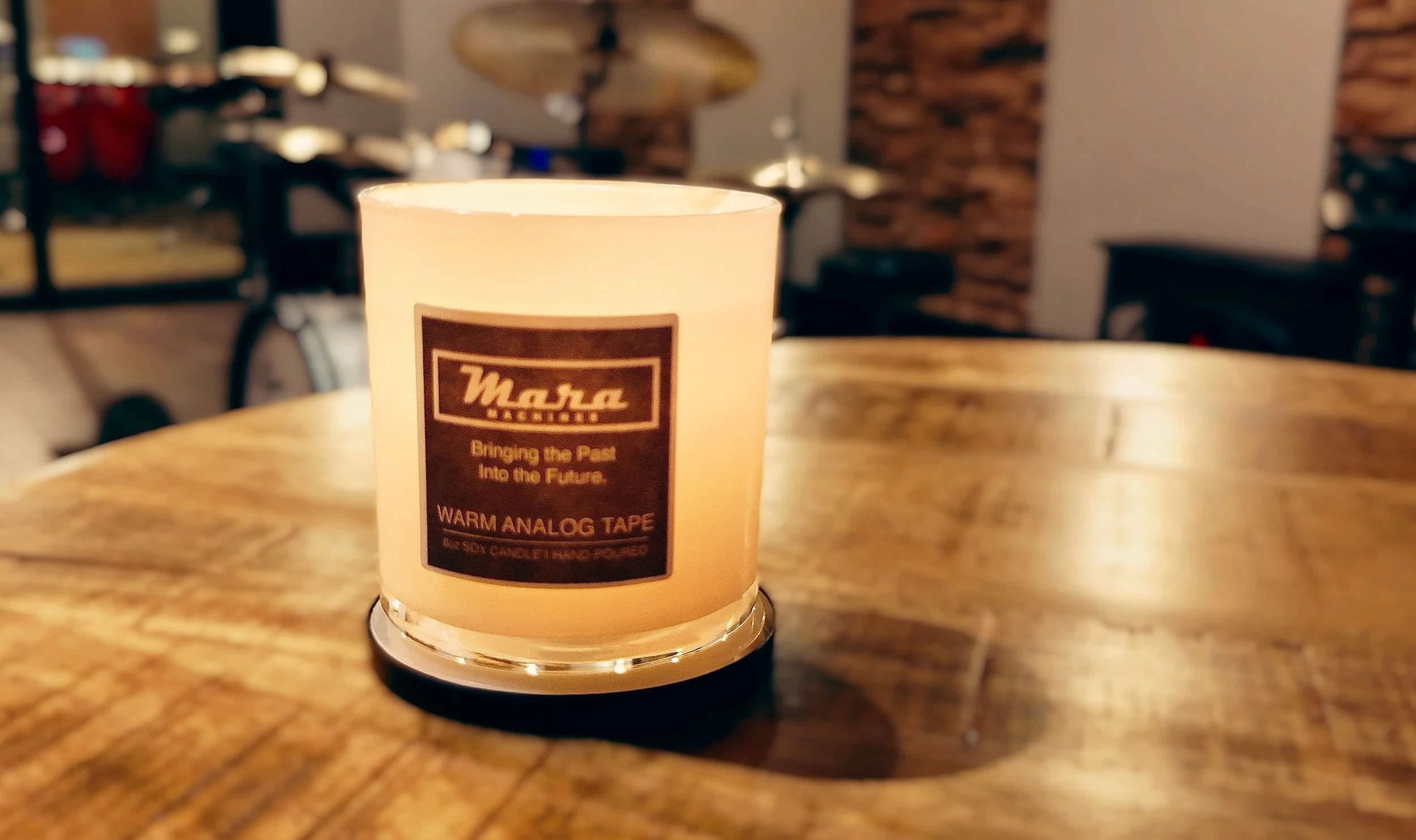 A lit white candle with a label reading 'Masa', sitting on a wooden table in a cozy indoor setting with dim lighting and a blurred background featuring a bicycle and a brick accent wall.